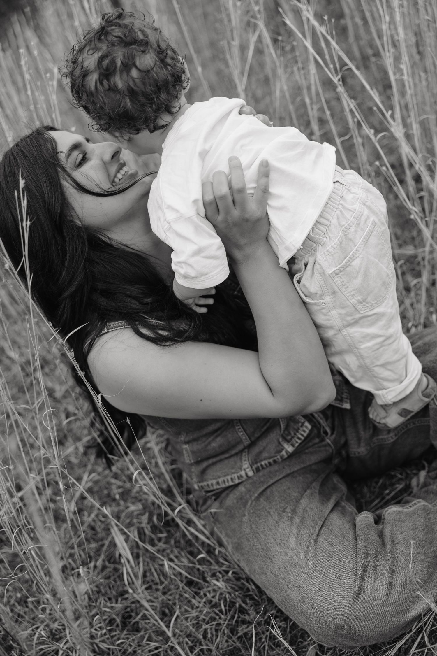 Black and white photo of a mother holding her young child in a field of tall grass, sharing a joyful moment.