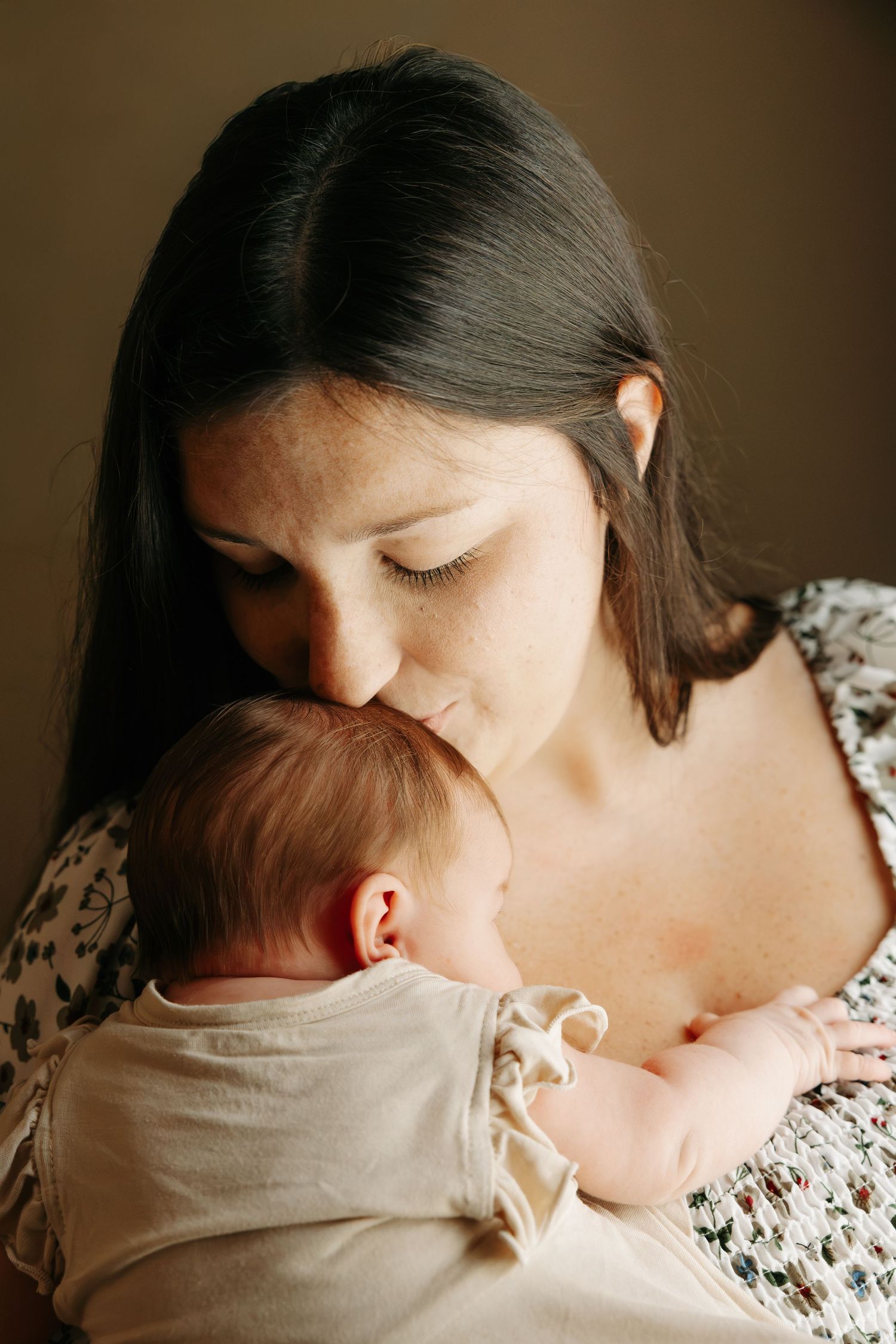 A mother with dark hair gently kisses her newborn baby's head while cradling the infant in her arms.