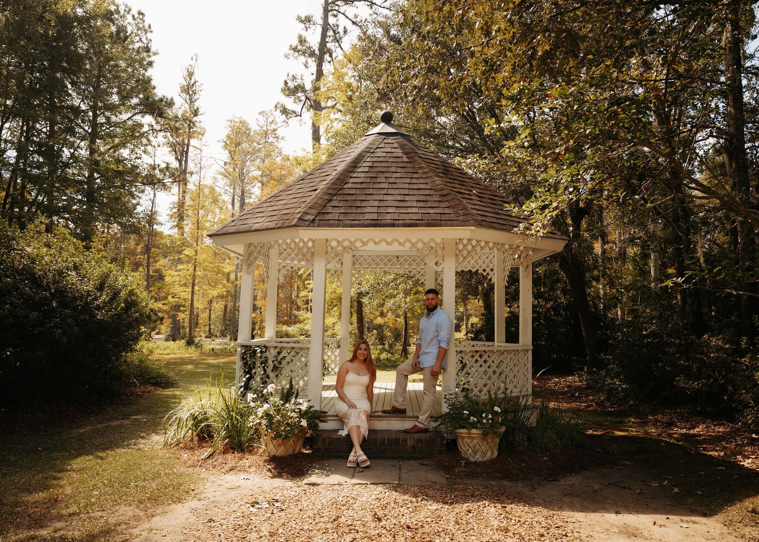 Couple standing together in a white gazebo surrounded by lush green trees and potted flowers in a garden setting.