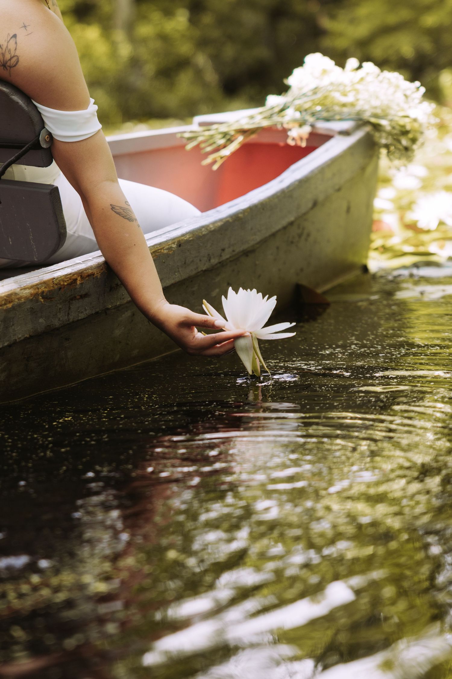 A woman in a boat gently places a white water lily on a calm pond, with flowers resting in the red-interior wooden boat.
