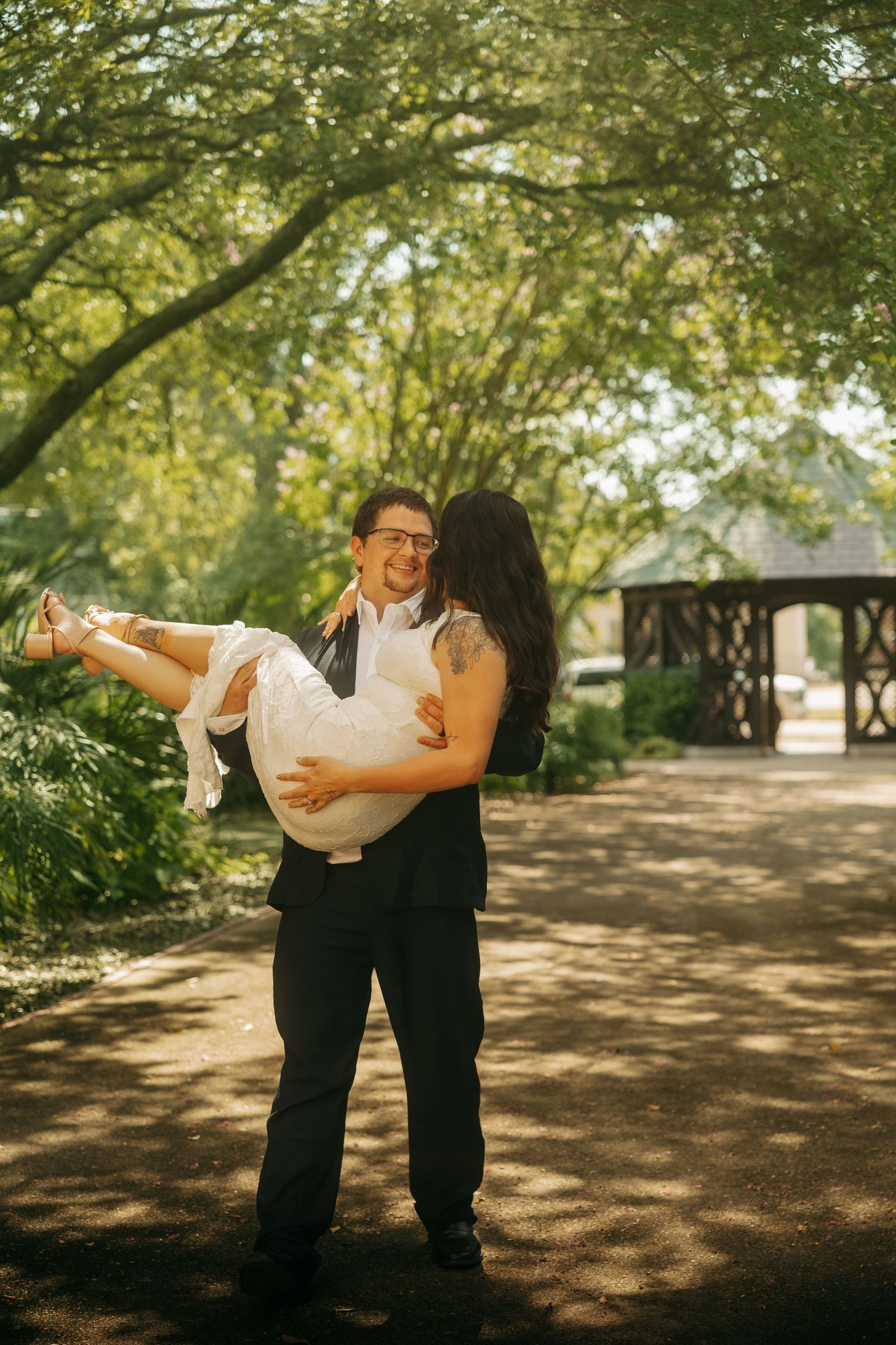 Groom carrying bride in white dress through a sunlit garden path with lush green trees and gazebo backdrop.