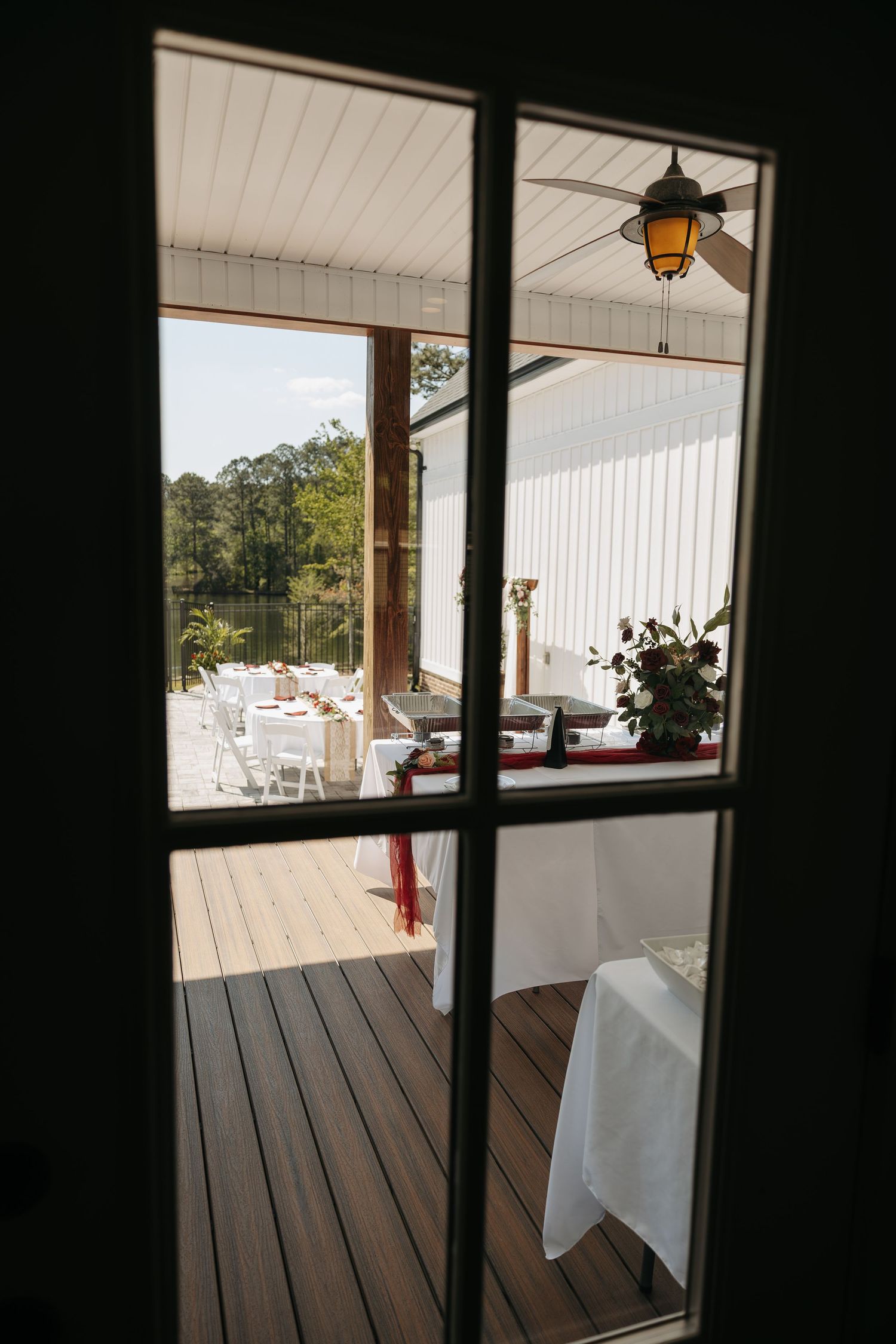 View through glass door of an outdoor wedding reception deck with white chairs and ceiling fan.