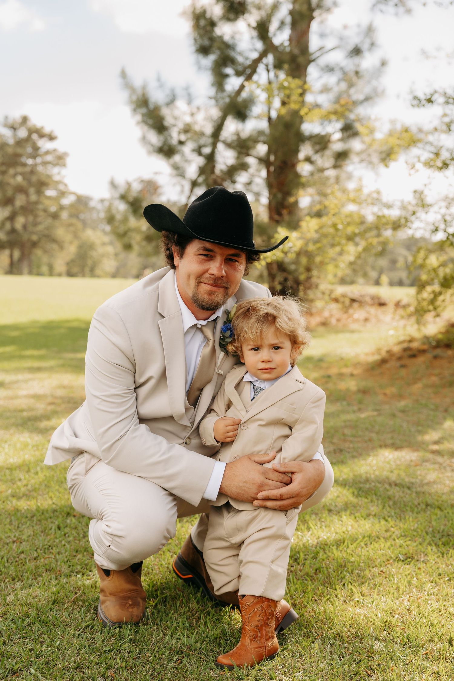 Groom in tan suit and black cowboy hat crouches beside a young boy in matching attire outdoors.