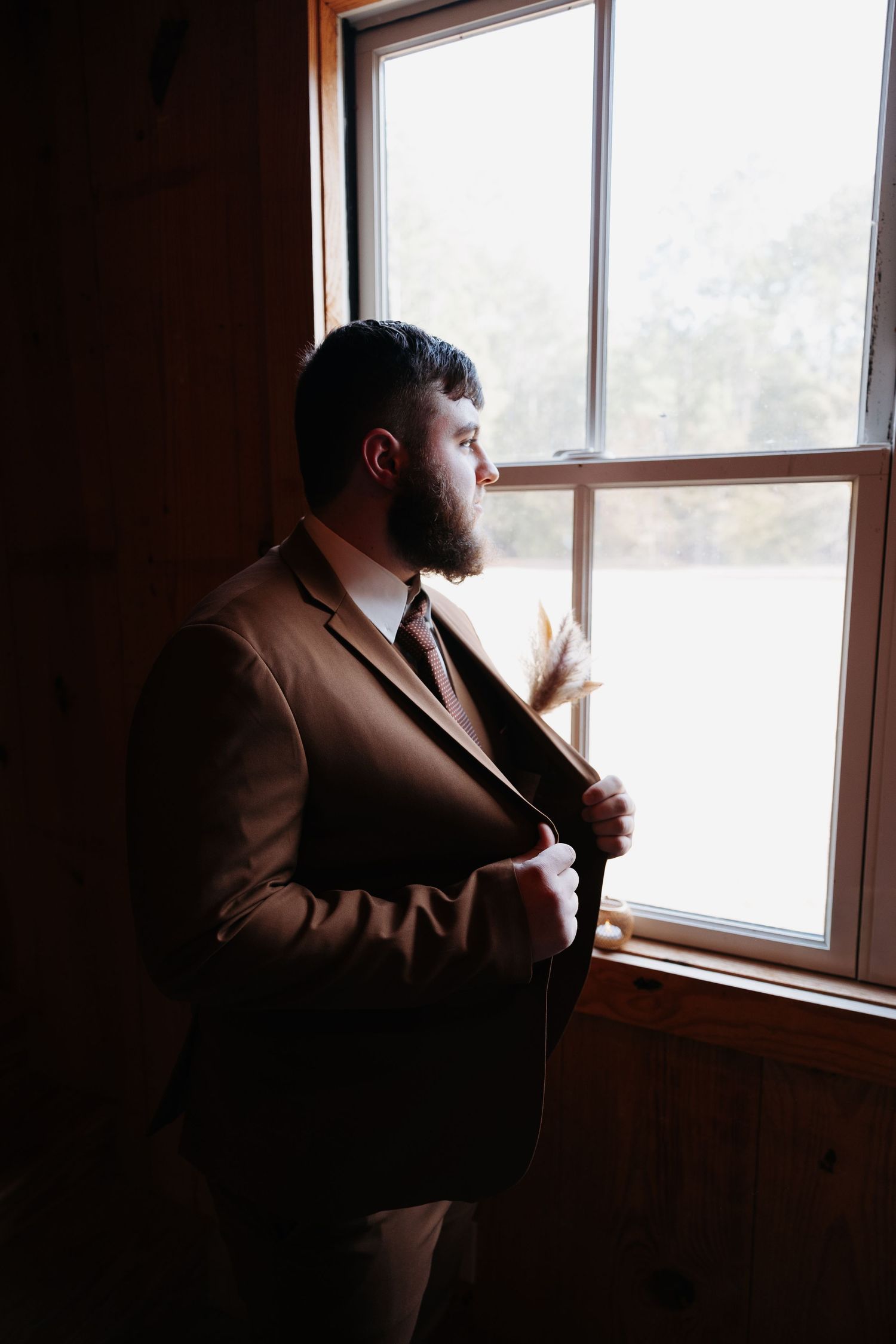 Bearded man in brown suit adjusting tie while gazing out a wooden-framed window in soft natural light.