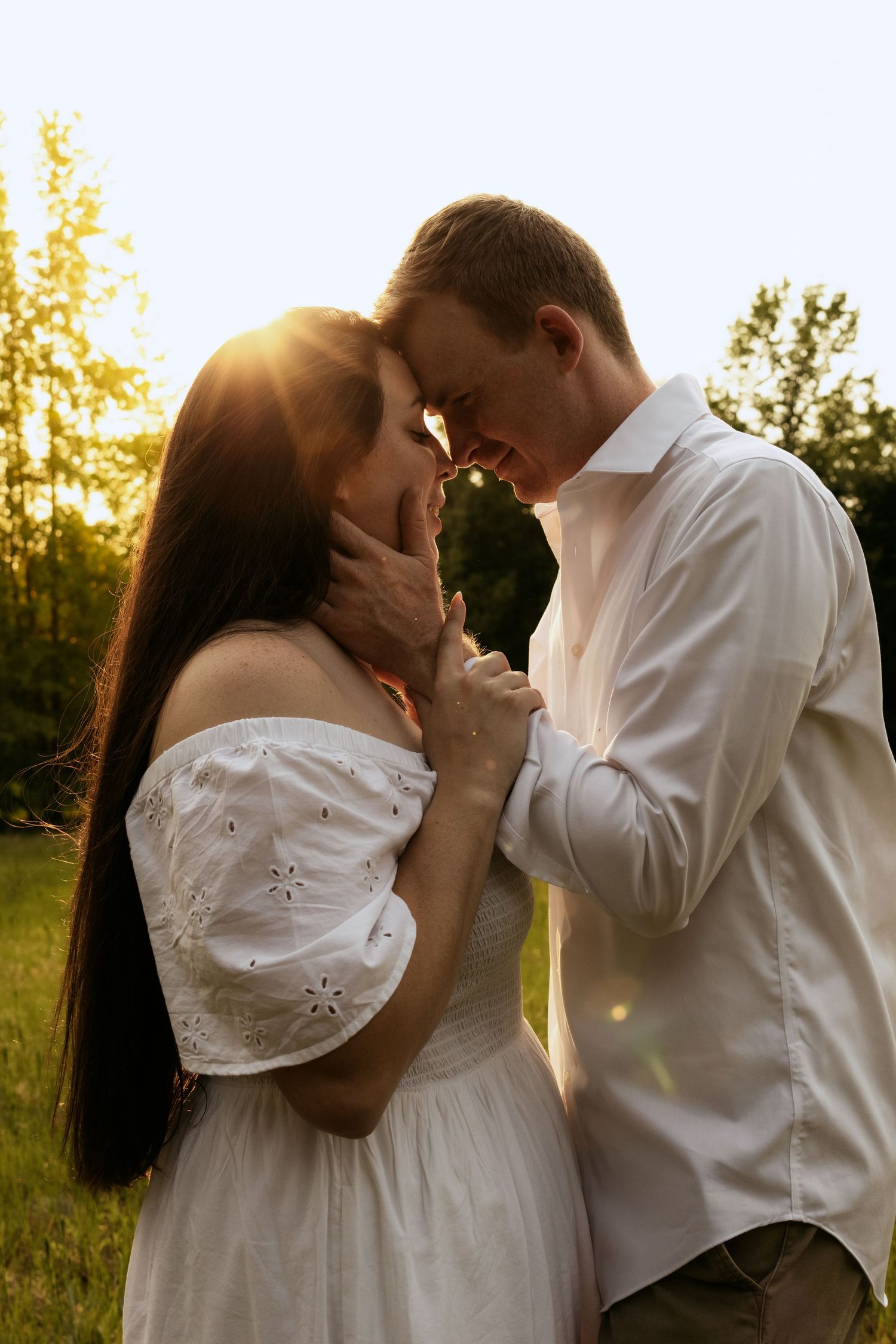 Romantic couple in white outfits sharing a tender moment at golden sunset in a lush green park.