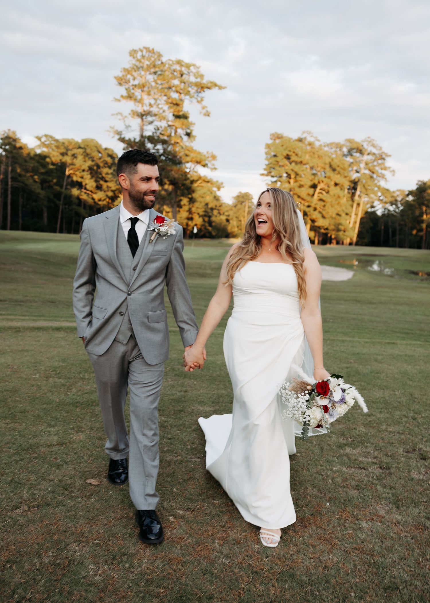 Happy bride and groom holding hands walking across a golf course on their wedding day at sunset.
