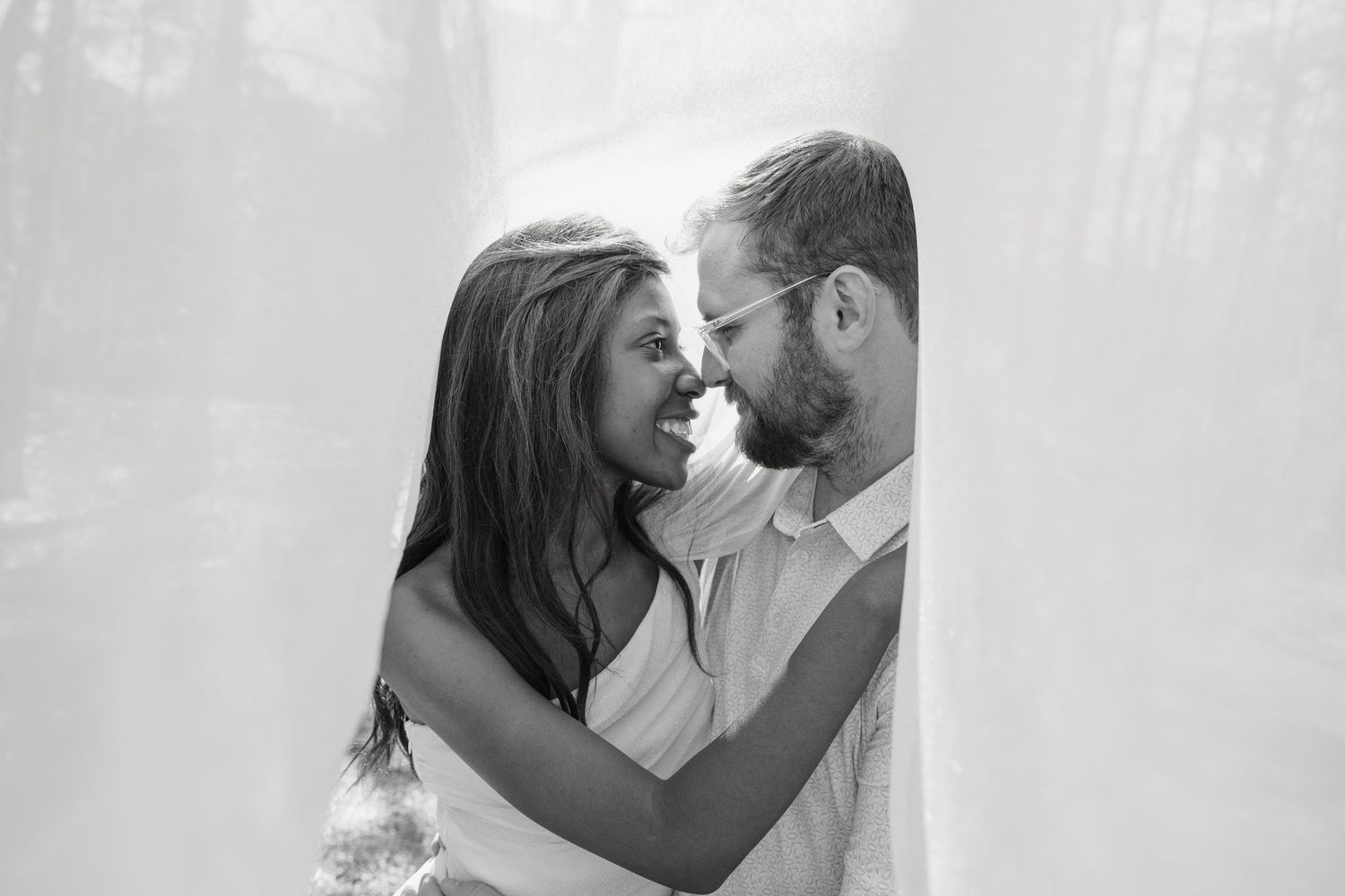 Black and white photo of a couple smiling and embracing intimately, framed by sheer white curtains.