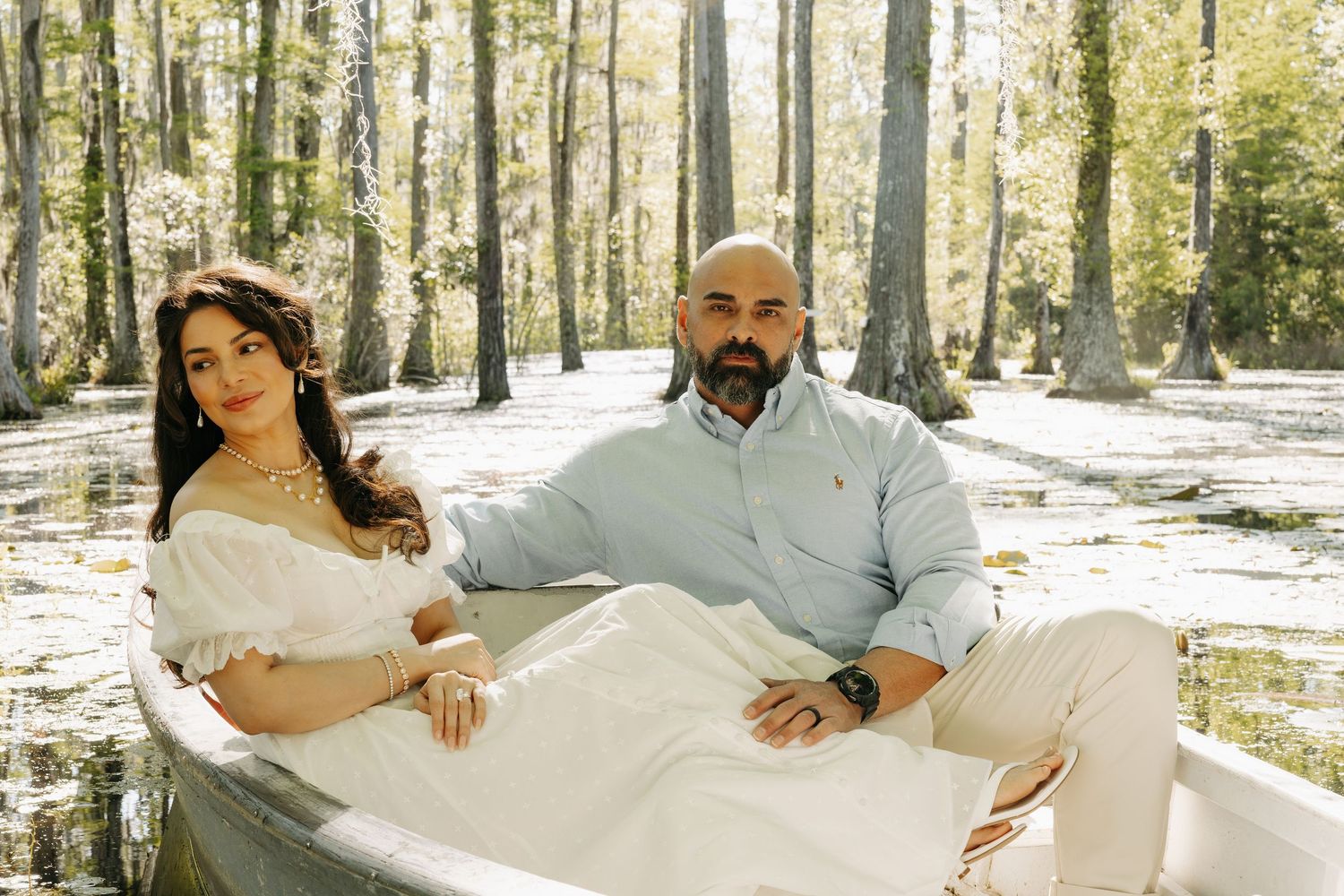 A couple poses romantically in a white boat surrounded by cypress trees in a serene swamp setting.