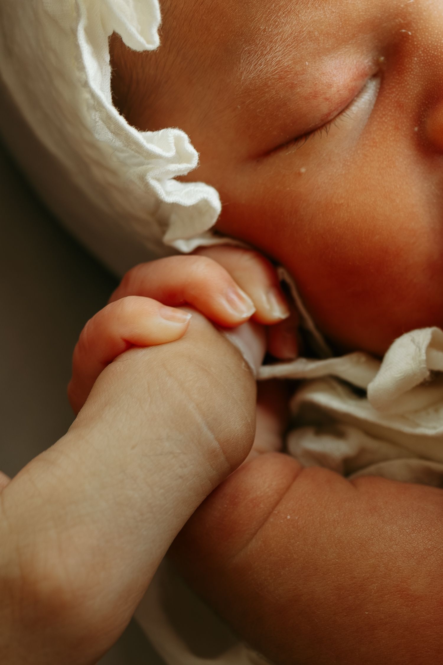 Sleeping newborn baby in white headband gently holds an adult finger, showcasing tiny delicate hands in a tender moment.