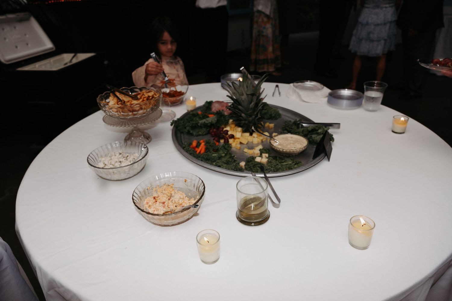 Elegant wedding appetizer table with charcuterie, fruit, and candles on a white linen-covered round table.