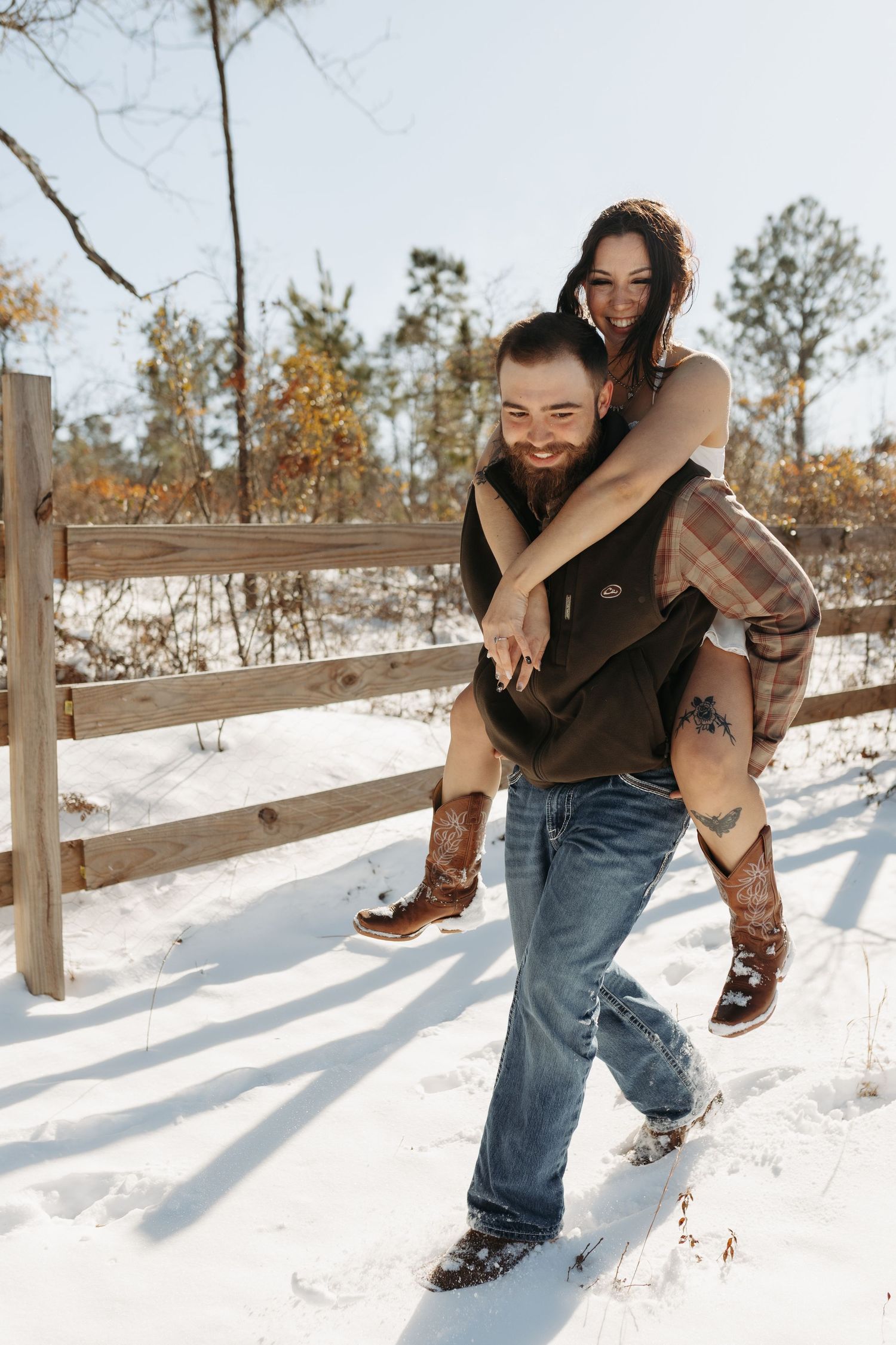 A bearded man gives a smiling woman a piggyback ride in the snow near a wooden fence in winter.