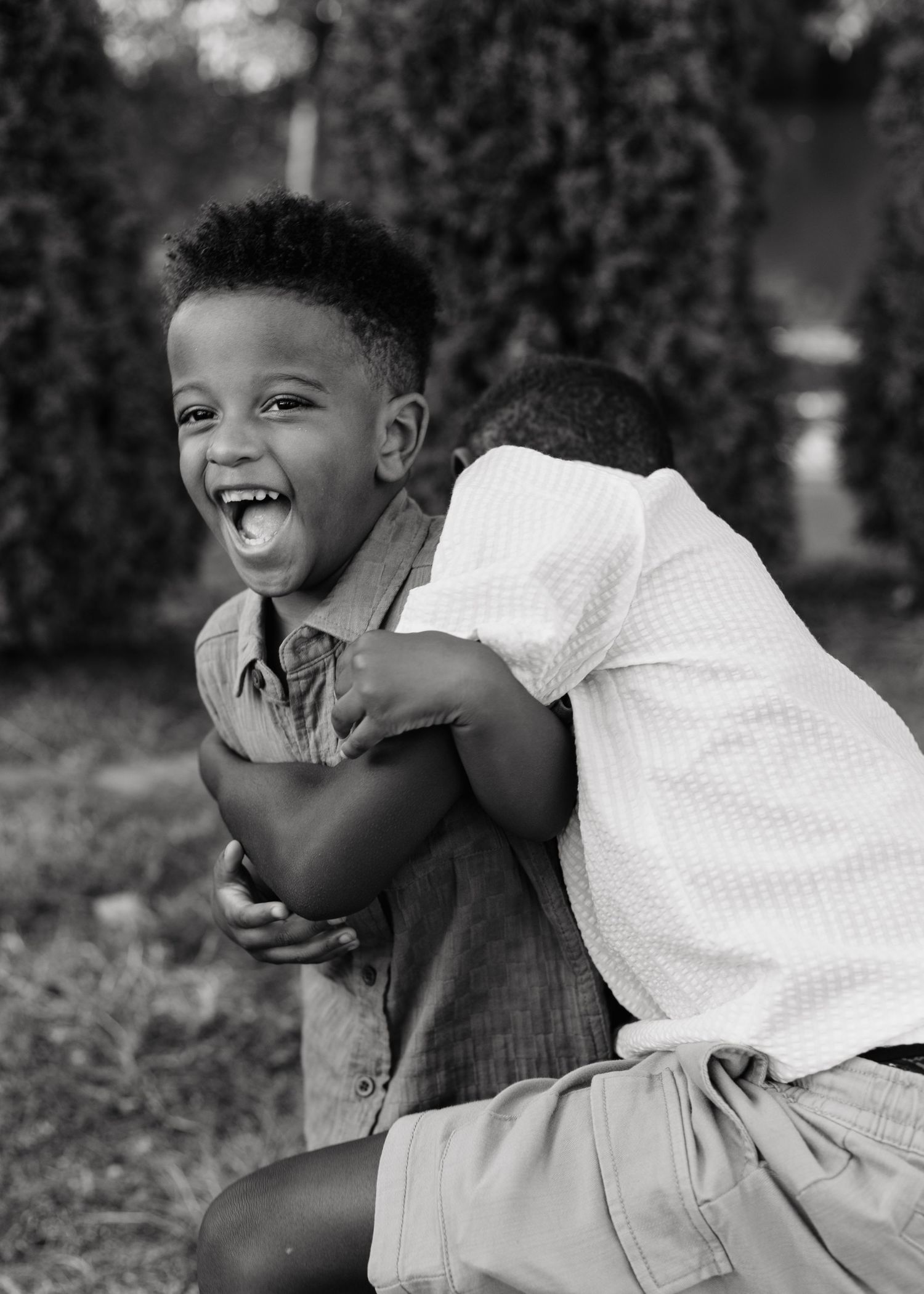 Two young boys share a joyful hug outdoors, one laughing brightly, captured in black and white photography.