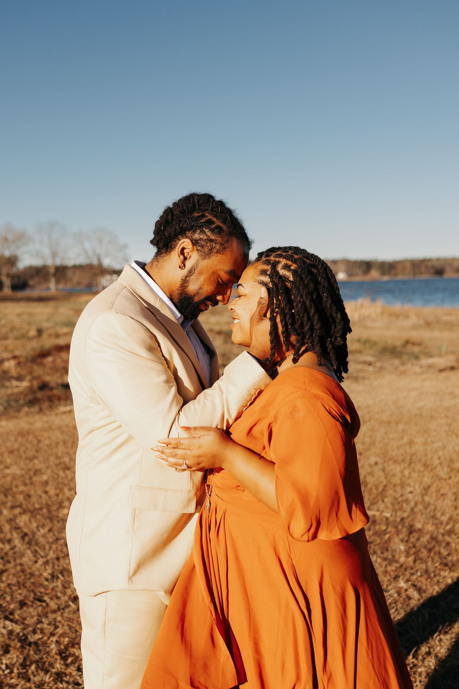 A couple in elegant attire shares a romantic moment outdoors near a lake on a sunny day.