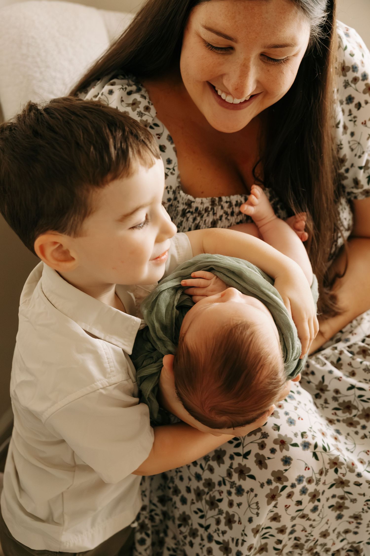 A smiling mother in a floral dress holds her newborn baby while her young son gazes lovingly at the infant.