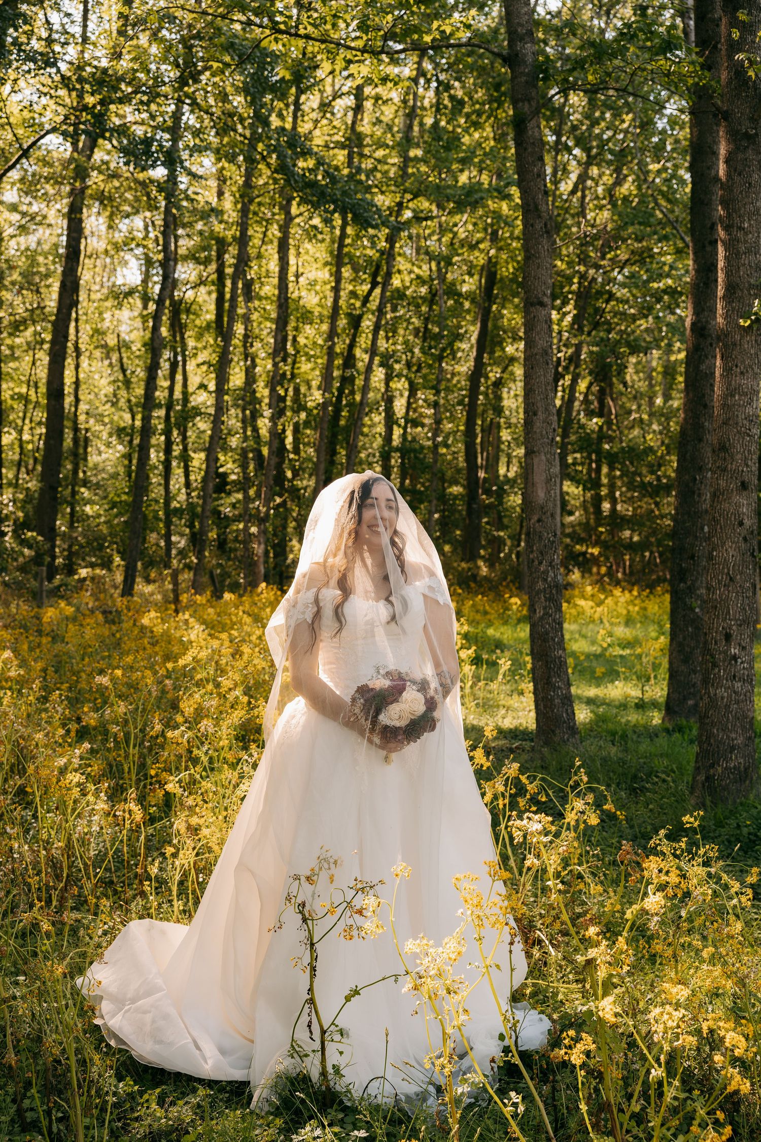 Bride in white ballgown and veil holds bouquet while standing among wildflowers in sunlit forest.