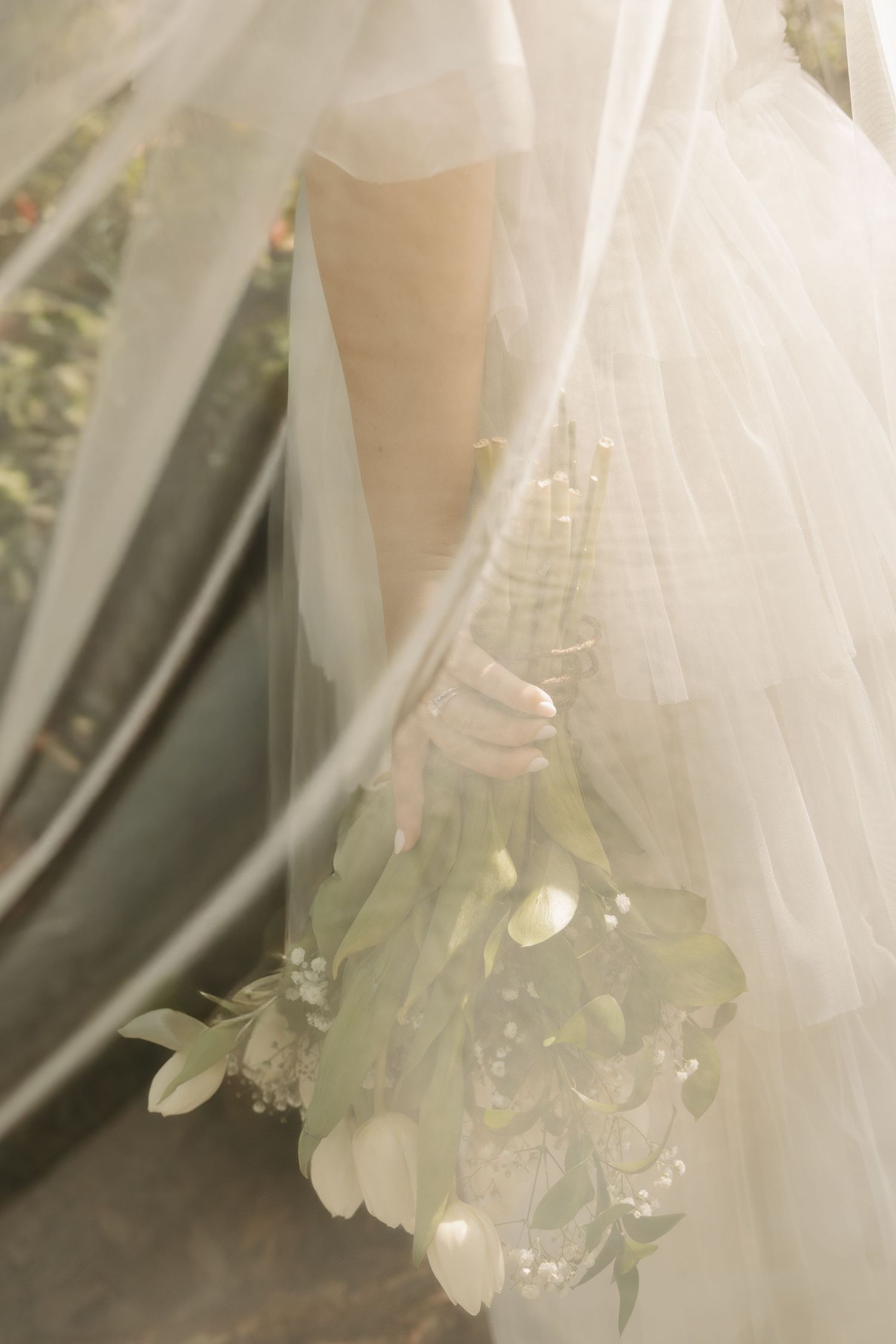 Bride holding a white tulip bouquet with greenery, partially veiled in soft sheer fabric on her wedding day.