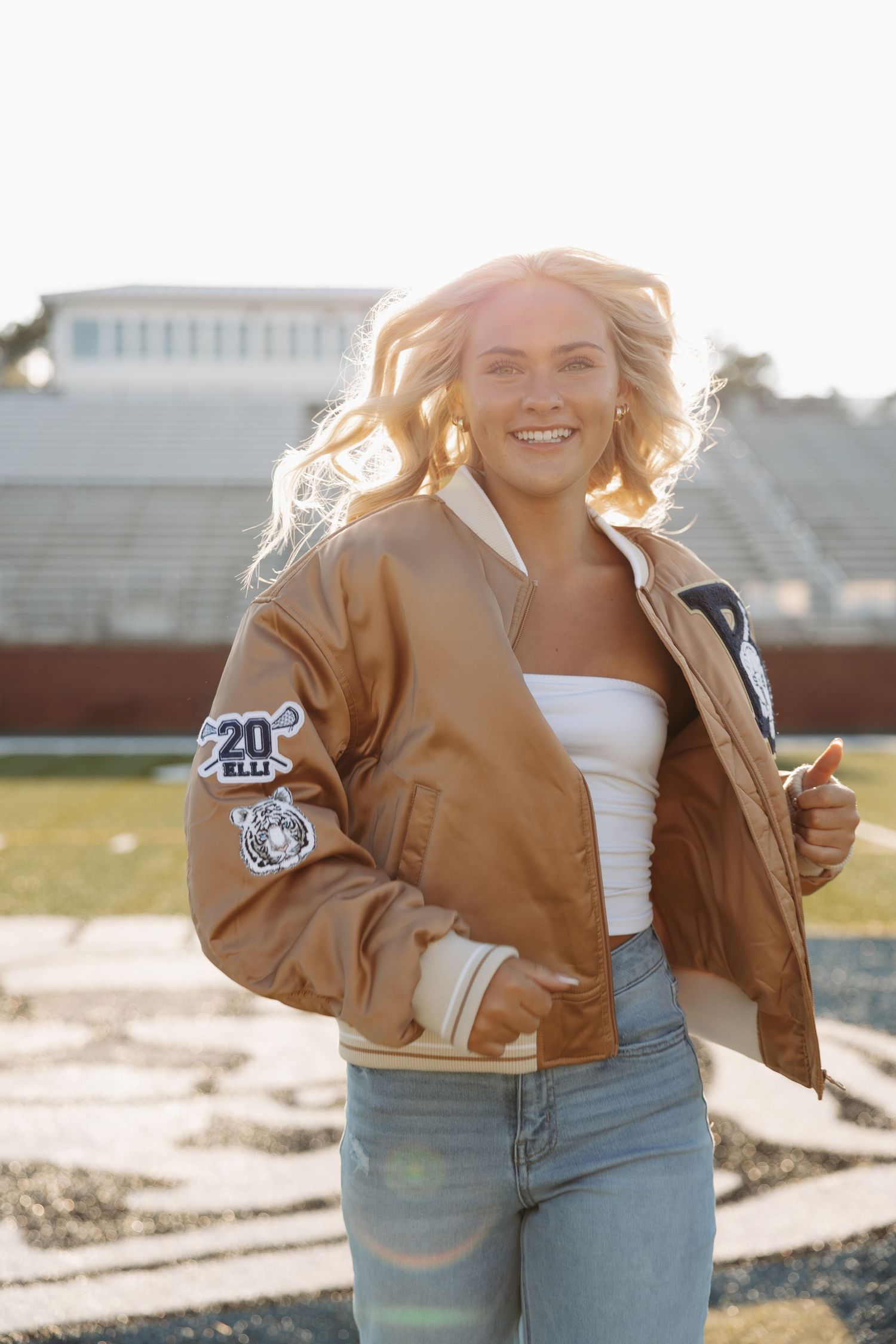 Light illuminates a casual outdoor scene with someone wearing a tan varsity jacket and jeans at a stadium.