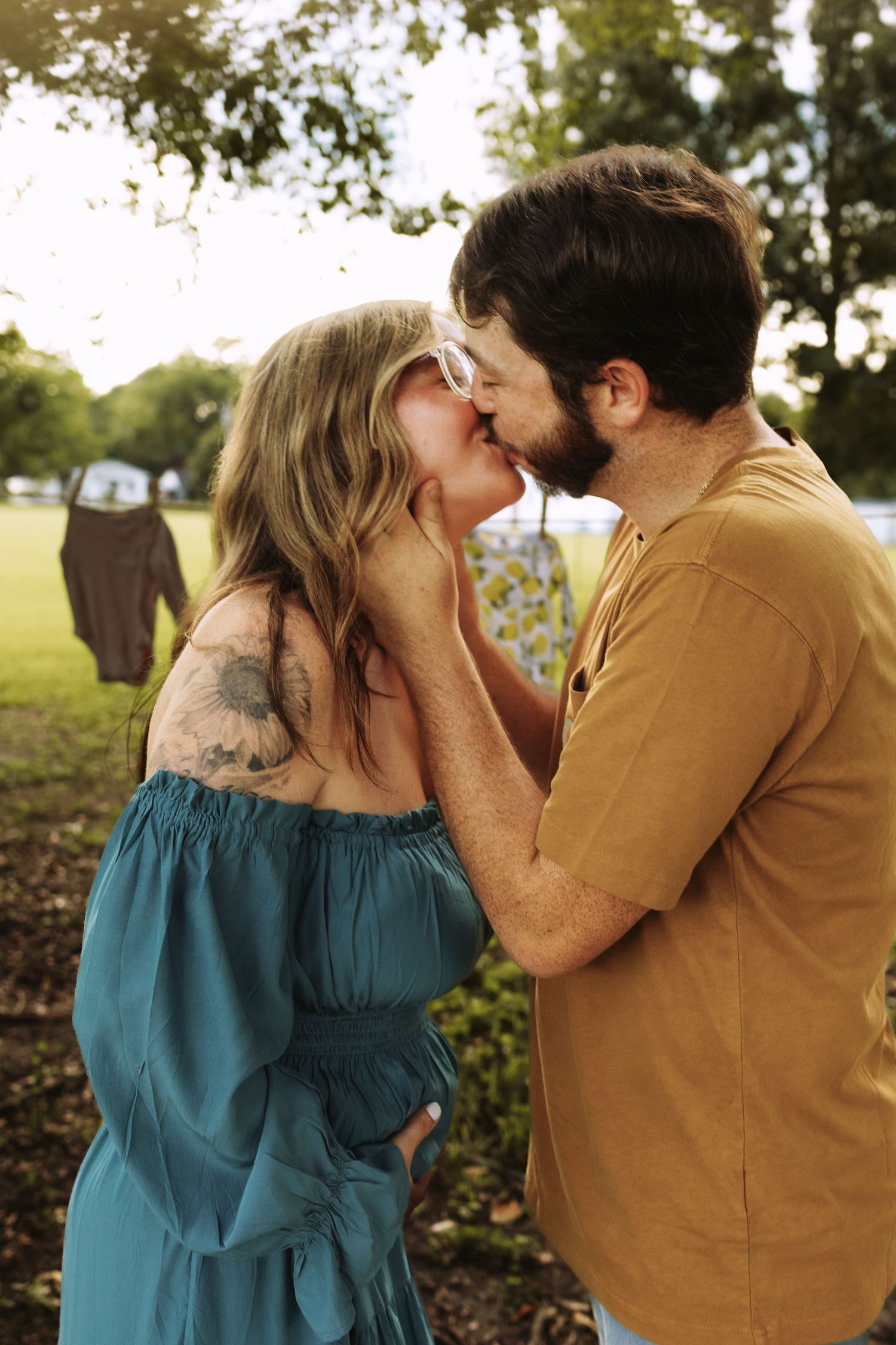 A couple wearing turquoise and mustard yellow outfits share a romantic kiss in a sunlit outdoor setting.