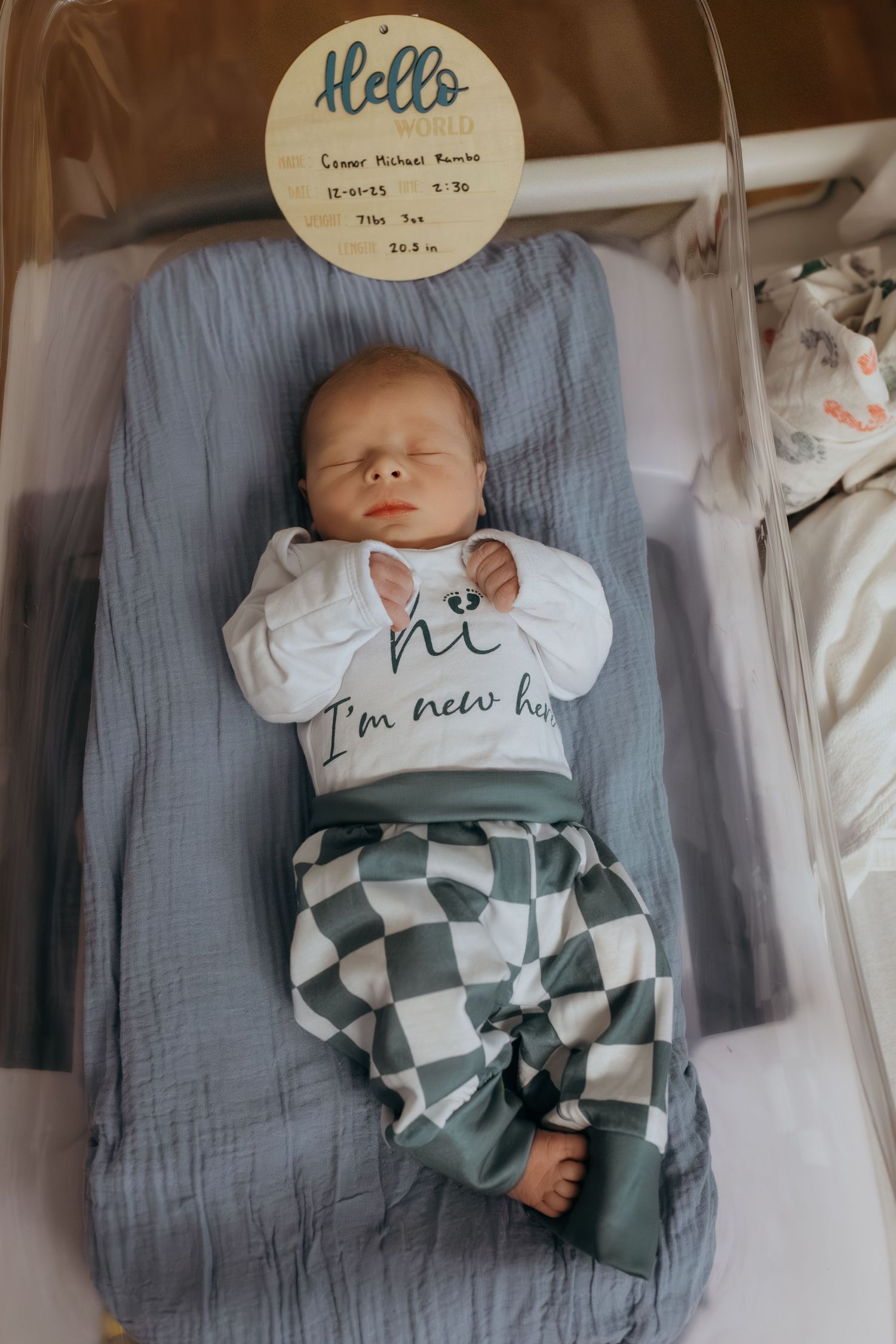 Newborn baby sleeping peacefully on blue blanket wearing checkered pants and 'I'm new here' shirt.