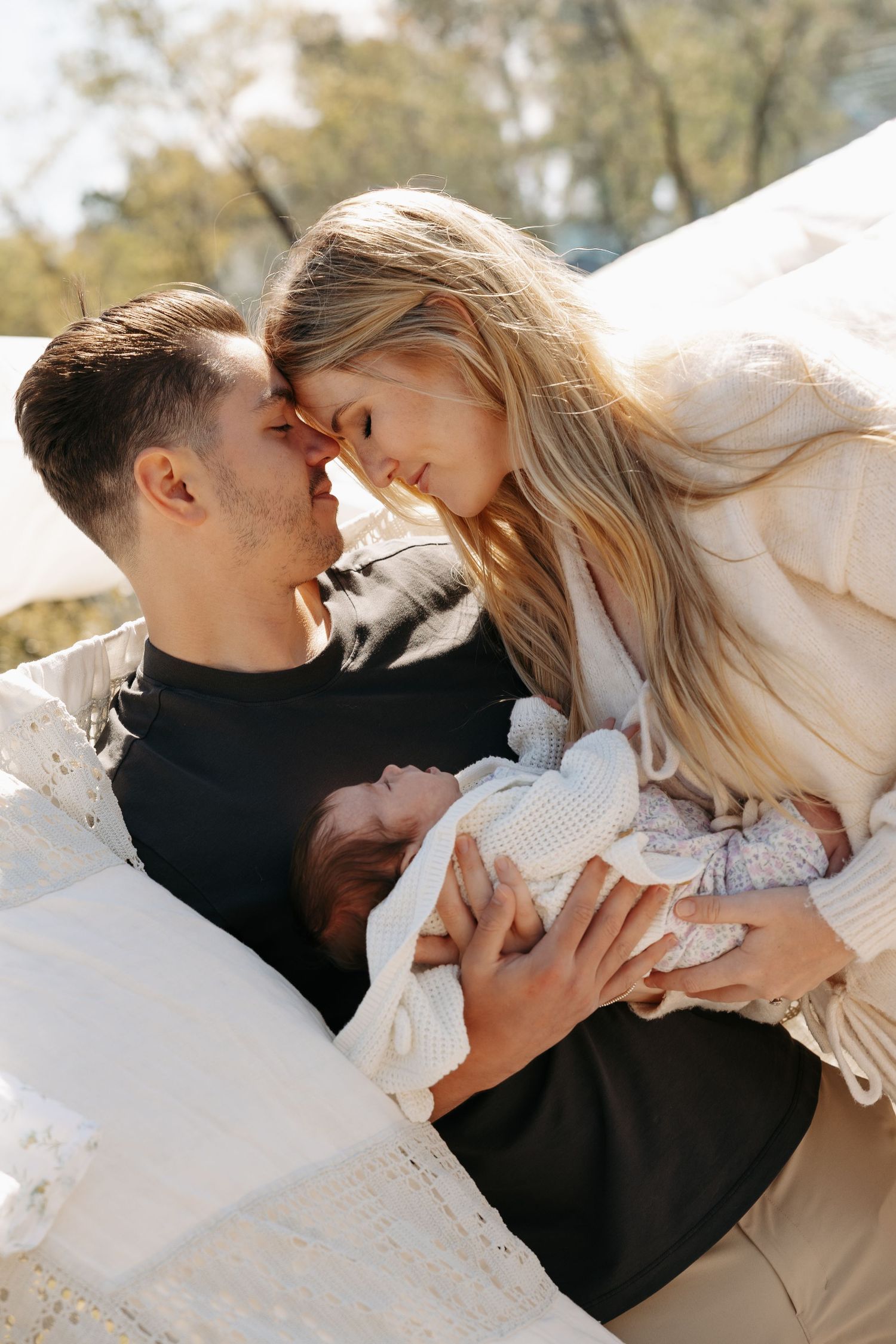 A young couple shares a tender moment, foreheads touching, while holding their newborn baby outdoors in warm sunlight.