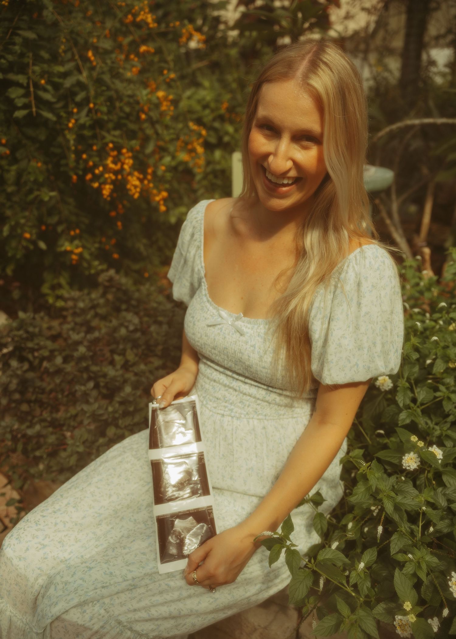 A figure in a vintage-inspired sage green dress poses with a small silver flask in a garden setting during autumn.