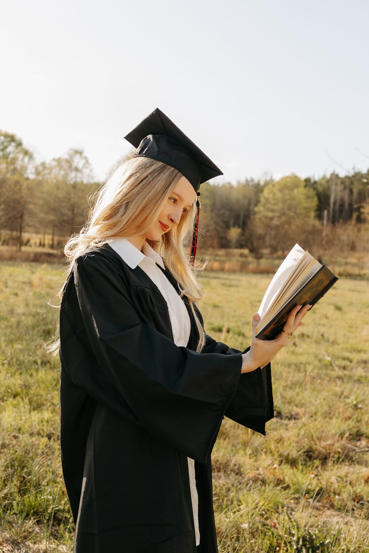 A blonde woman in a black graduation gown and cap reads an open book outdoors in a sunny green field.