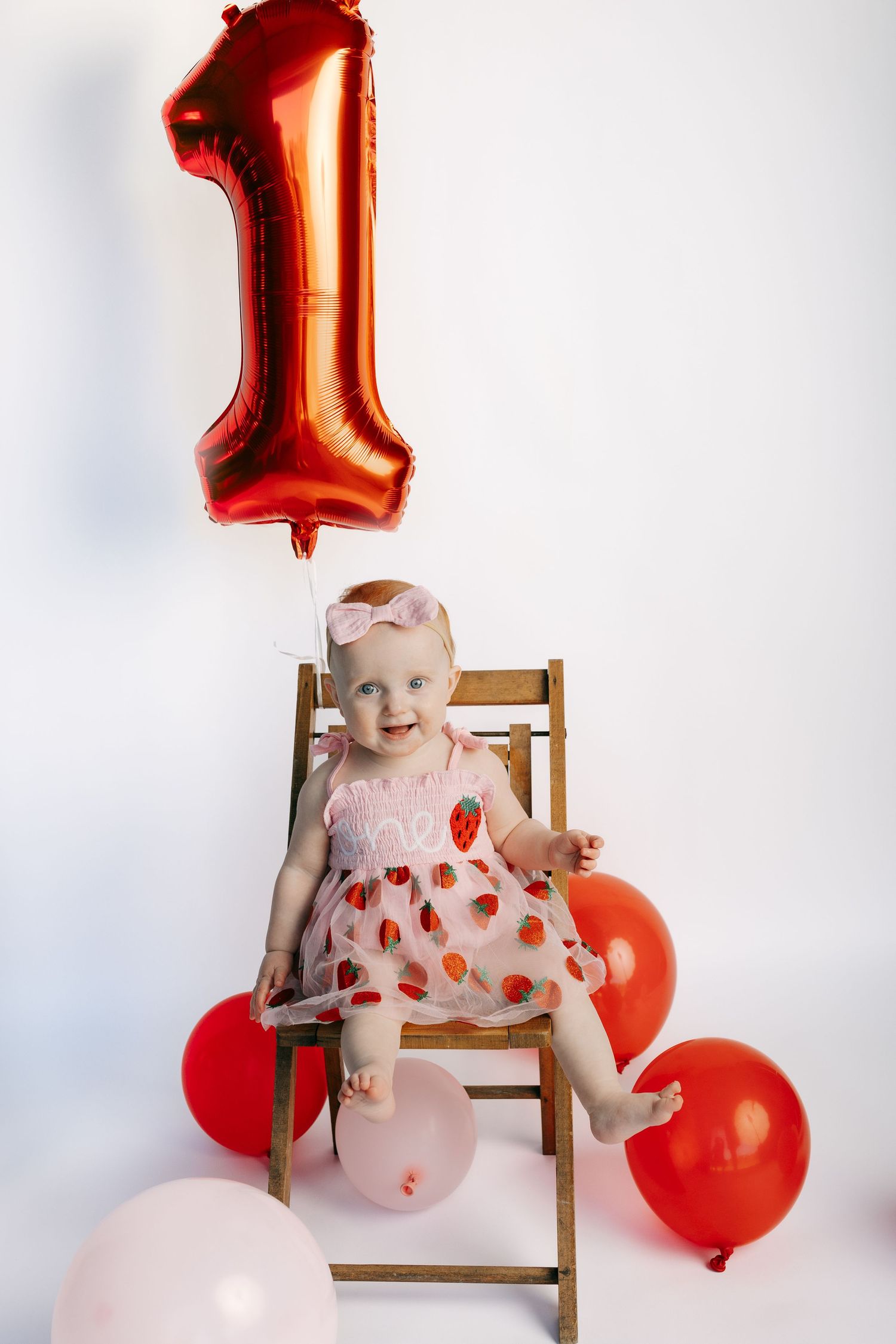 Baby girl in strawberry dress sits on wooden chair with red number 1 balloon celebrating her first birthday.
