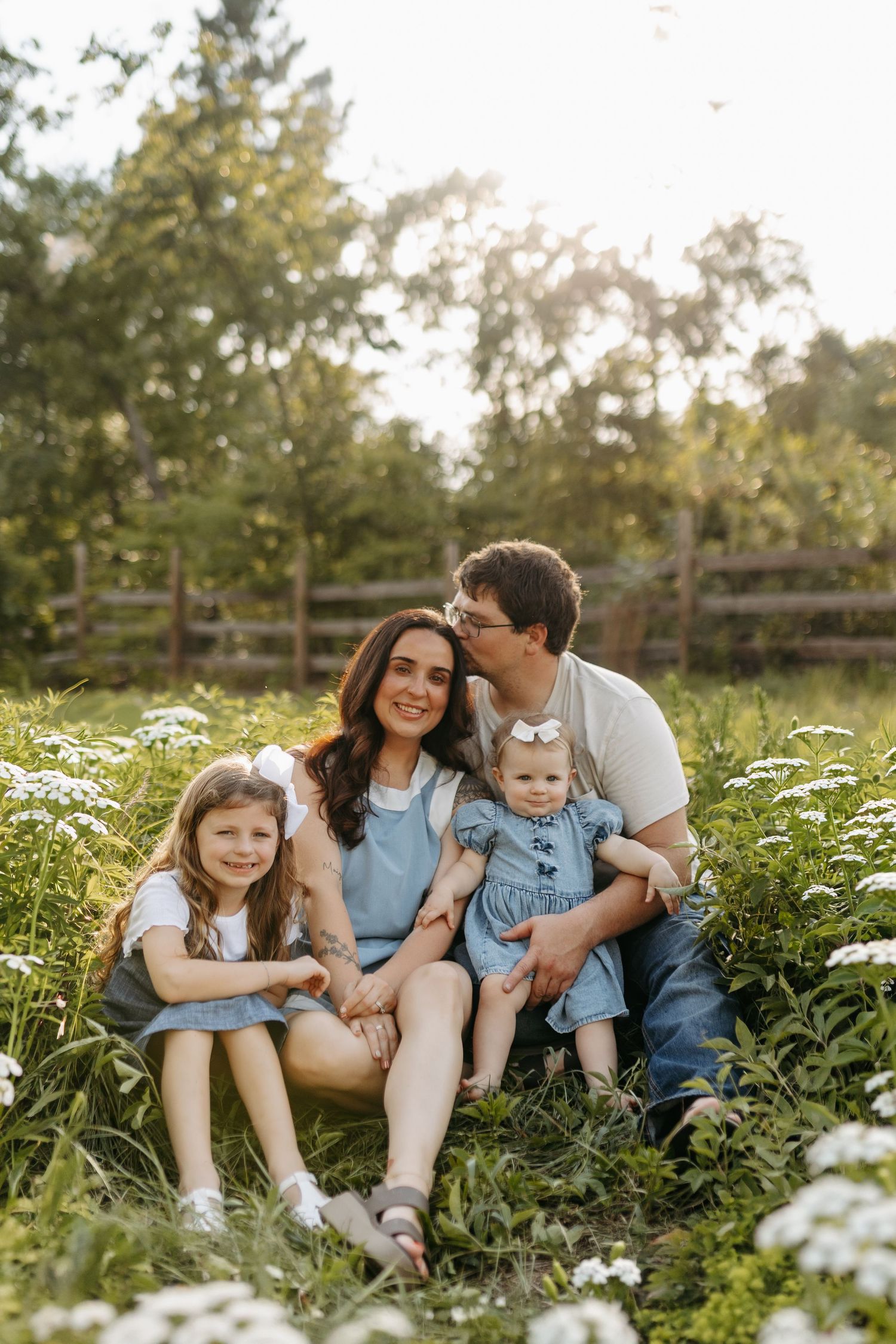 Happy family of four sitting in a sunny wildflower field, parents with two young daughters in matching denim outfits.