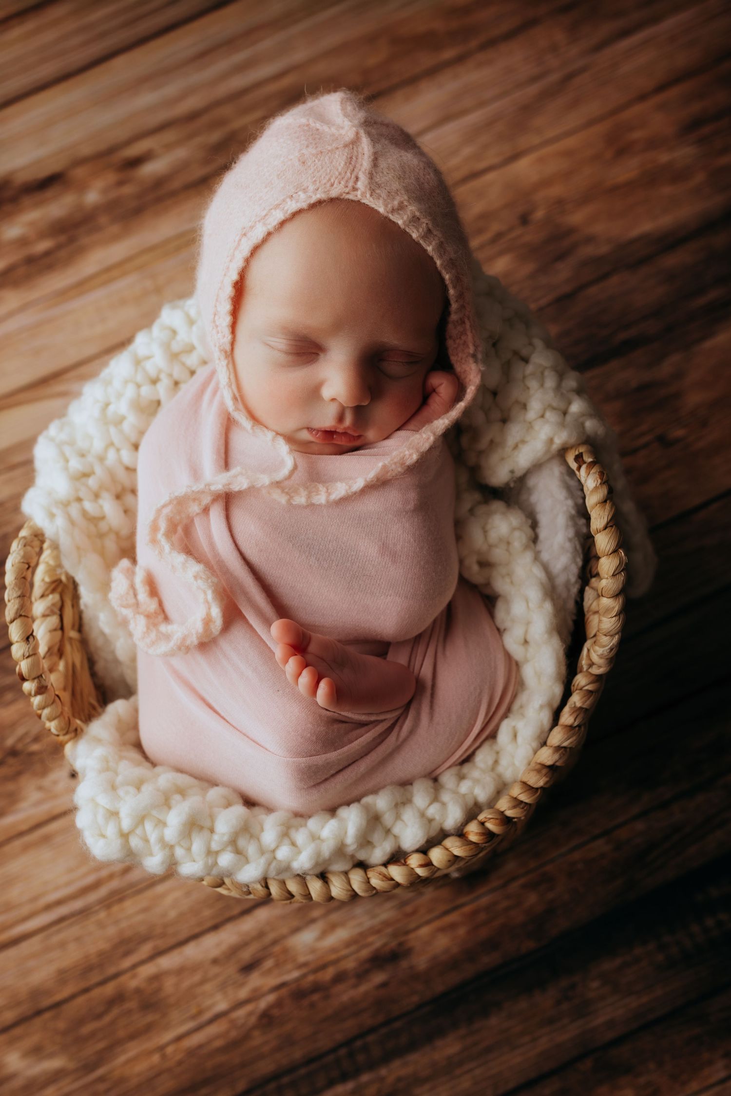 Newborn baby sleeping peacefully in a wicker basket, swaddled in pink wrap and bonnet on a wooden floor.