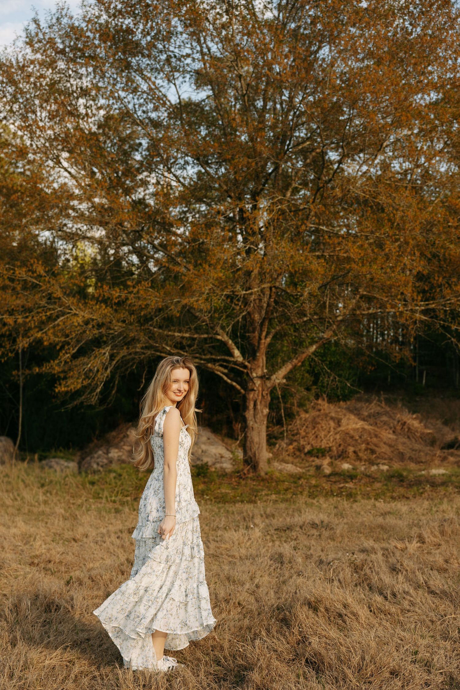 Woman in floral maxi dress standing in autumn field with golden trees in background.