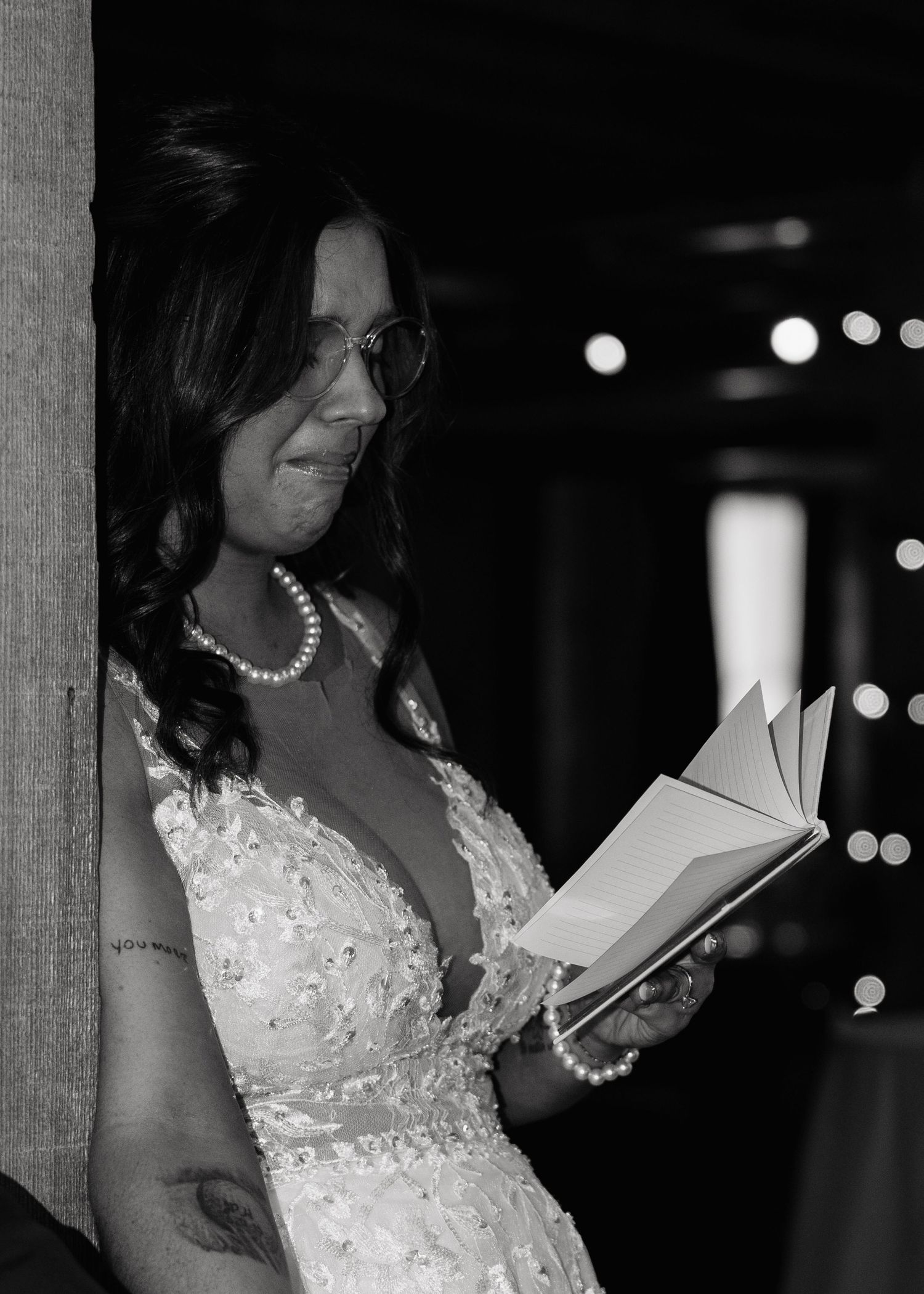 Bride in lace gown and pearl necklace reading vows from a book during wedding ceremony, black and white photo.