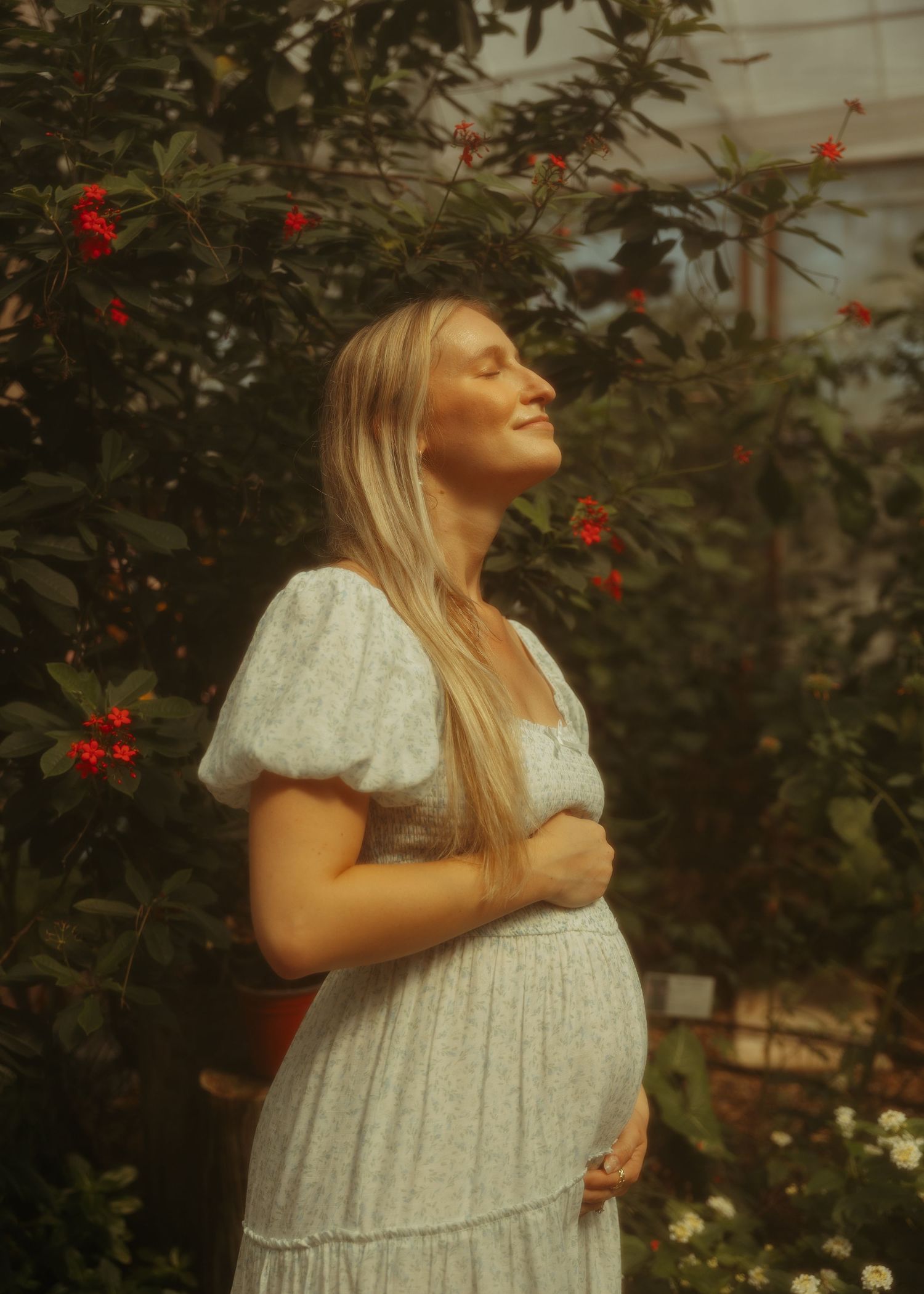 Pregnant woman in white dress smiling with eyes closed among red flowering plants in a greenhouse.