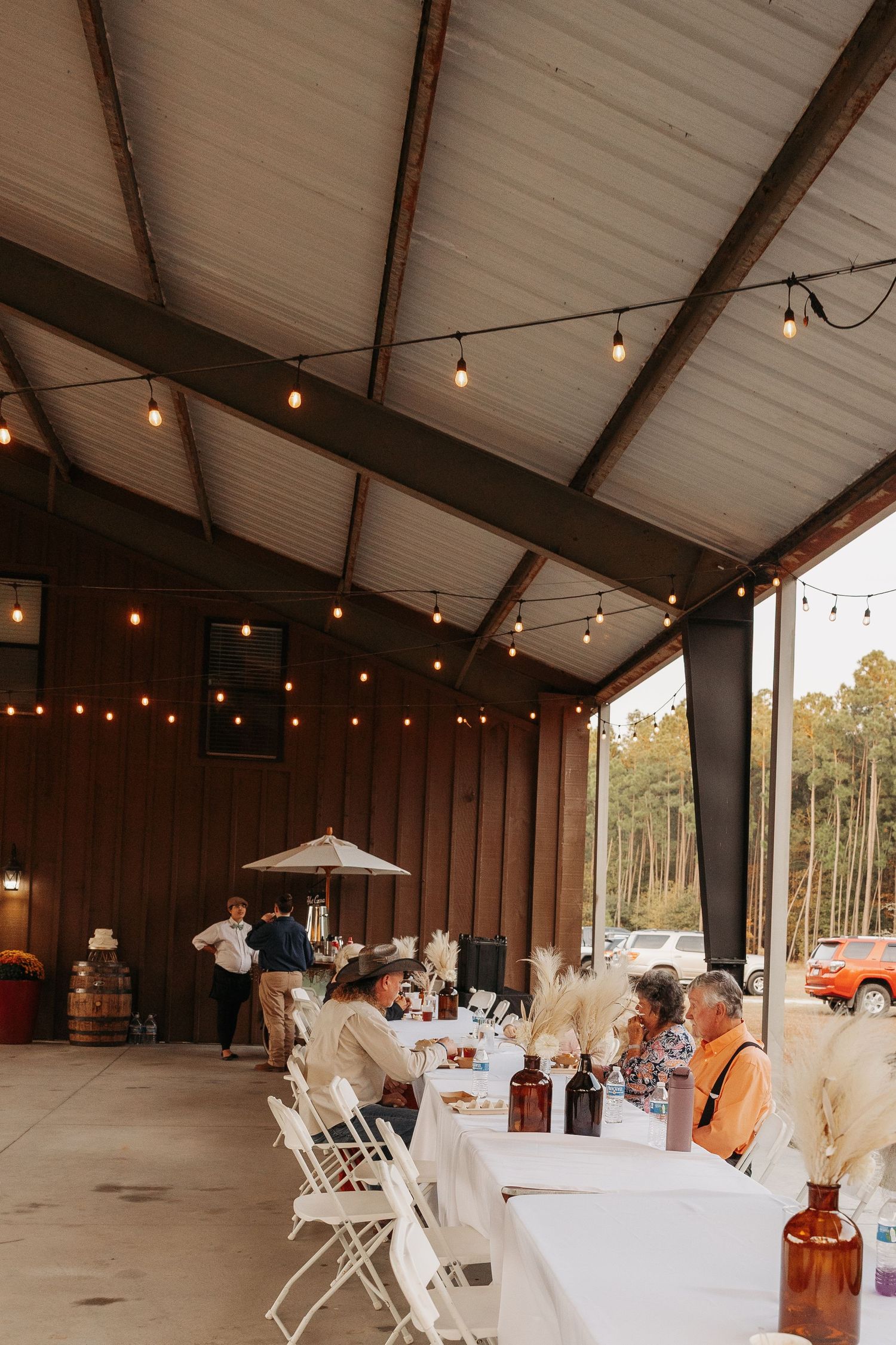 Rustic barn wedding reception with string lights, long white tables, and pampas grass centerpieces.