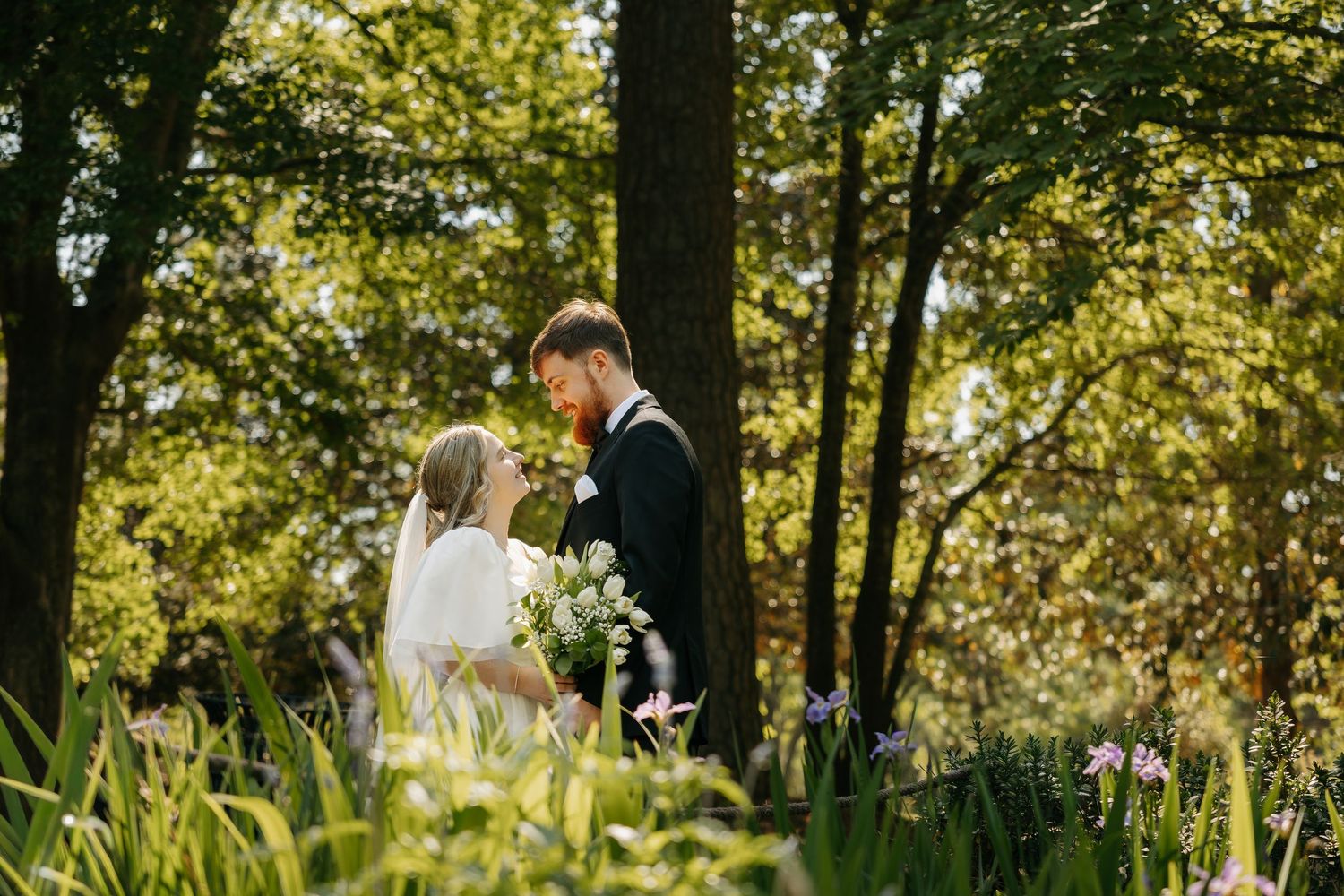 A bride and groom share a romantic moment surrounded by lush greenery and wildflowers in a sunlit woodland setting.