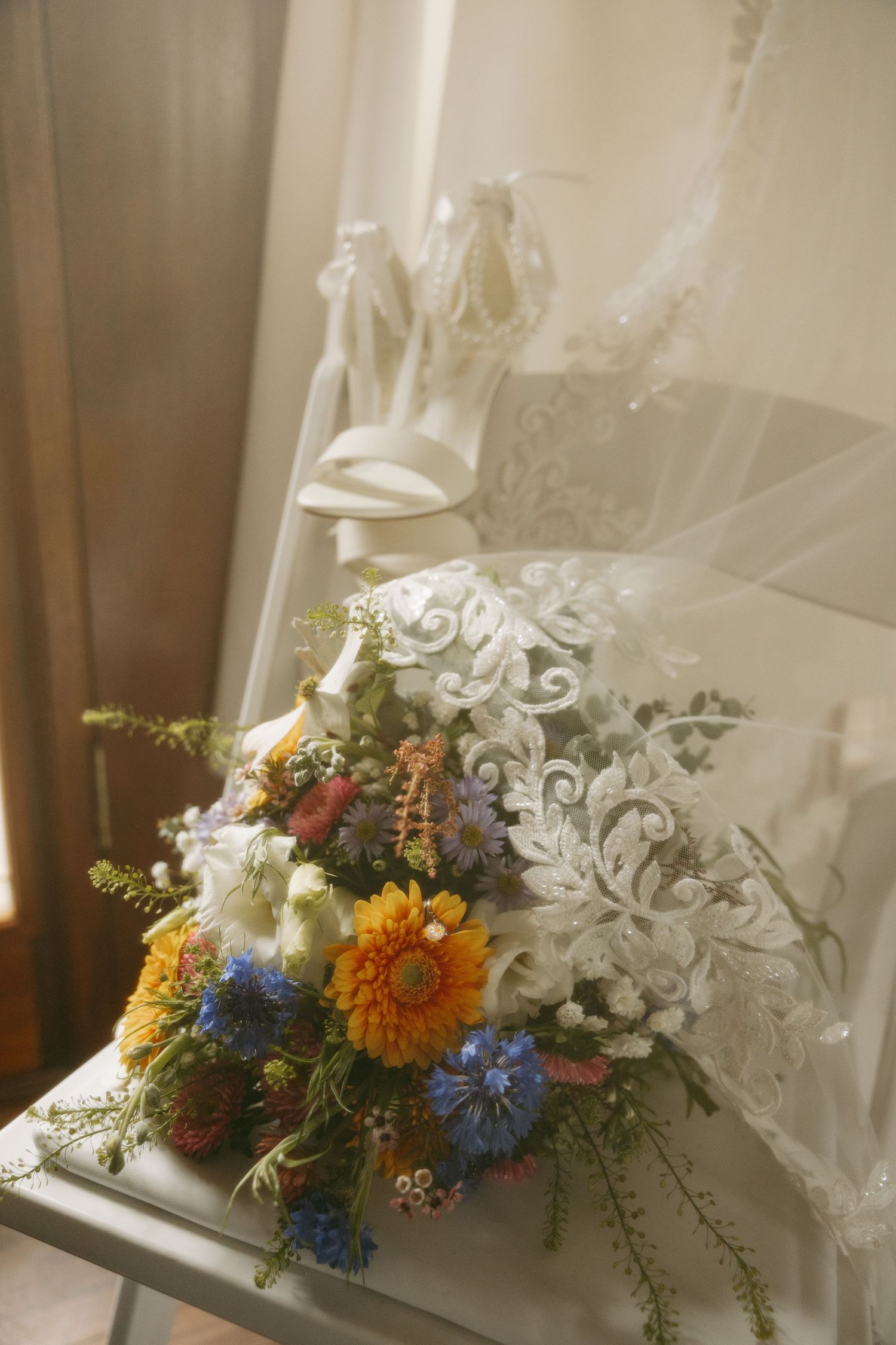 Colorful wildflower wedding bouquet with orange gerberas and blue flowers resting against a white lace wedding dress.