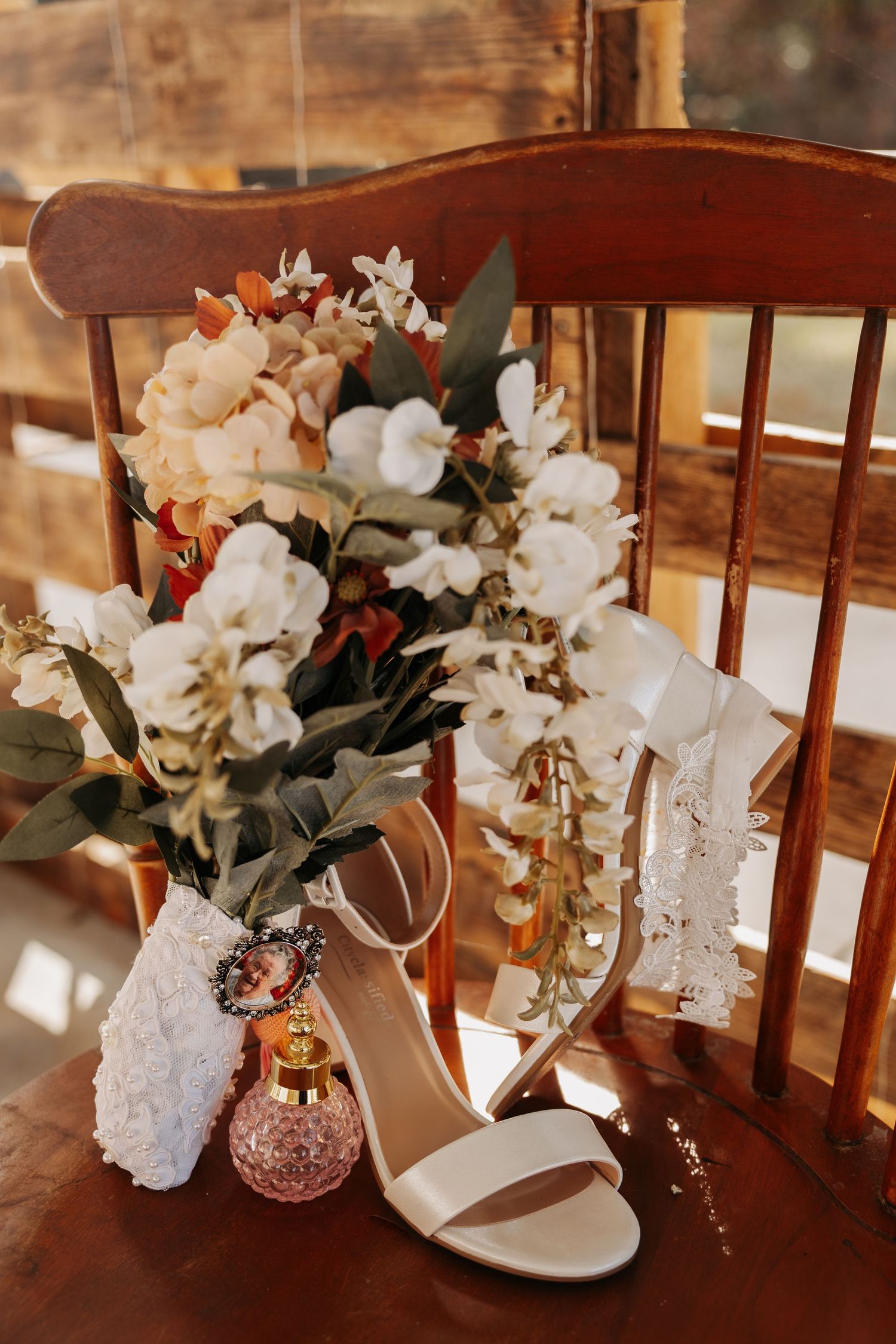 Bridal details flat lay featuring white heels, floral bouquet, perfume bottle, and lace accessories on a wooden chair.