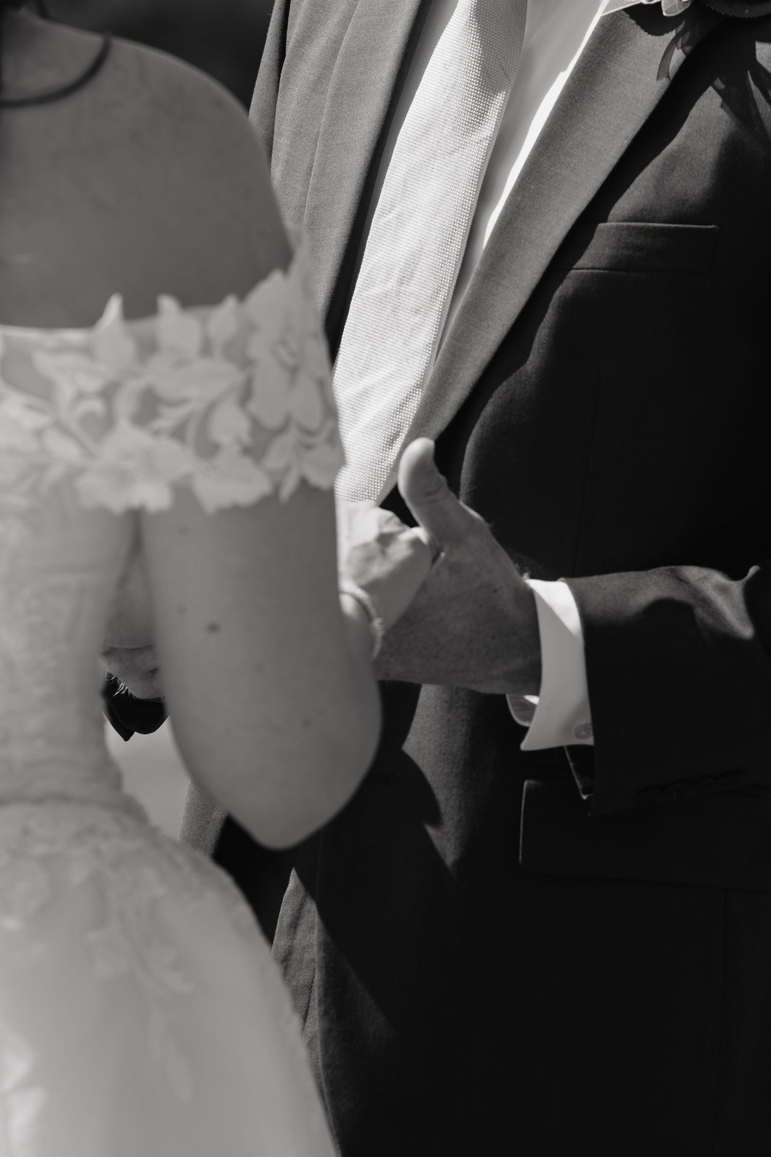 Black and white photo of bride and groom holding hands, showcasing lace wedding dress and dark suit.