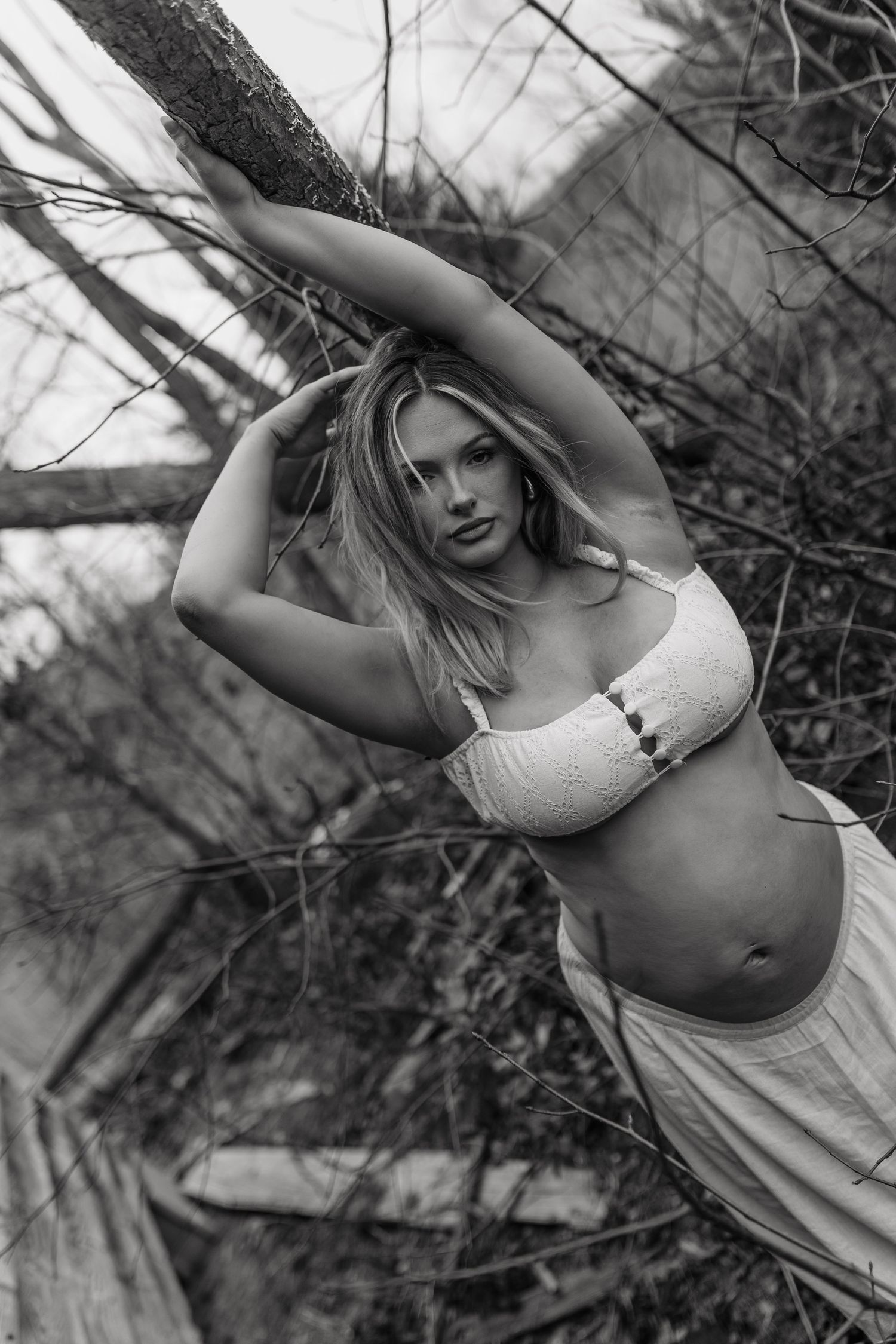 Black and white photo of a blonde woman in a white crop top and skirt posing among bare tree branches outdoors.