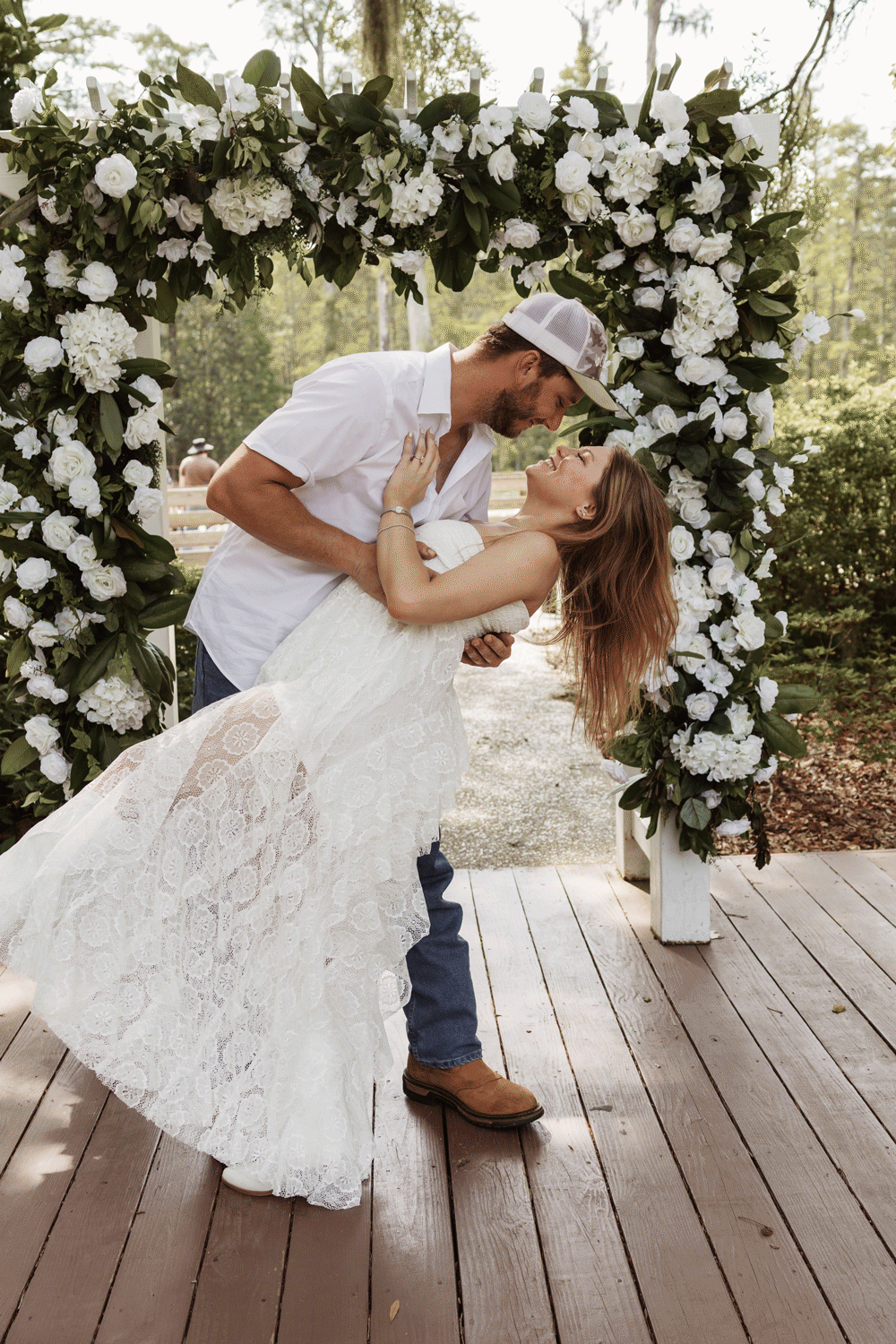 Couple sharing a romantic dip kiss under a white floral arch at an outdoor rustic wedding ceremony.