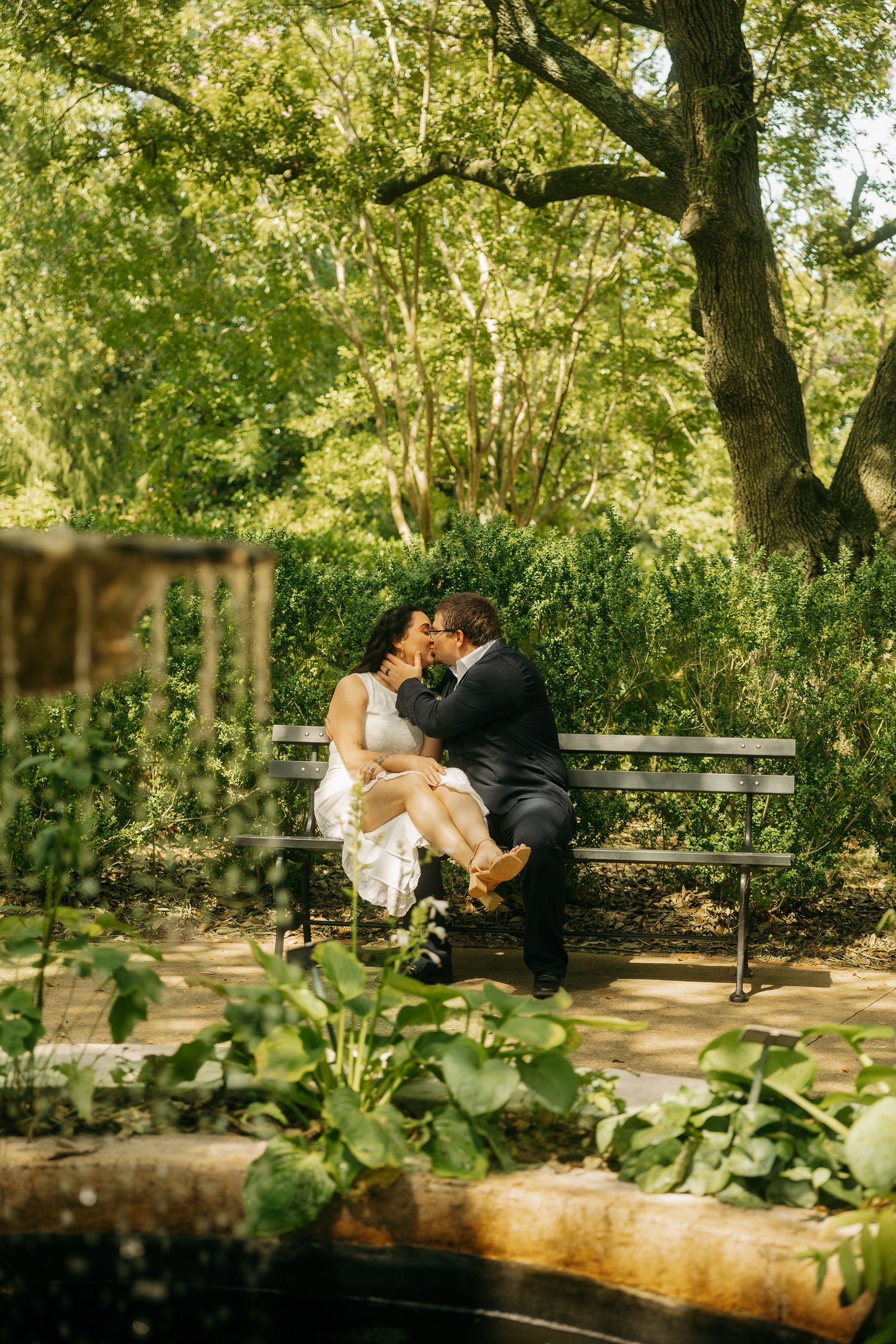 Bride and groom share a romantic kiss on a park bench surrounded by lush green trees and foliage.