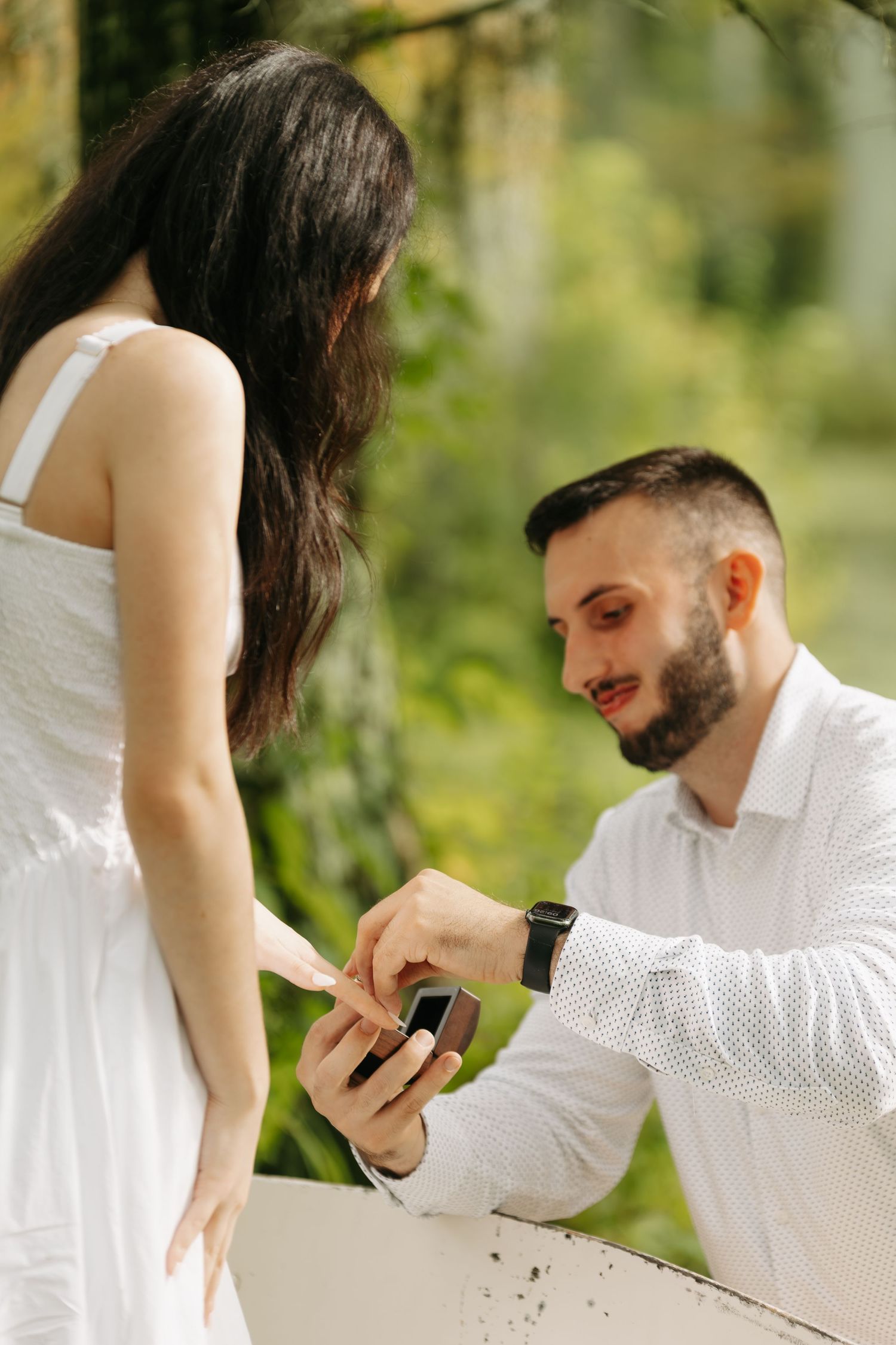 Man kneeling on one knee proposing to woman in white dress outdoors in lush green garden setting.