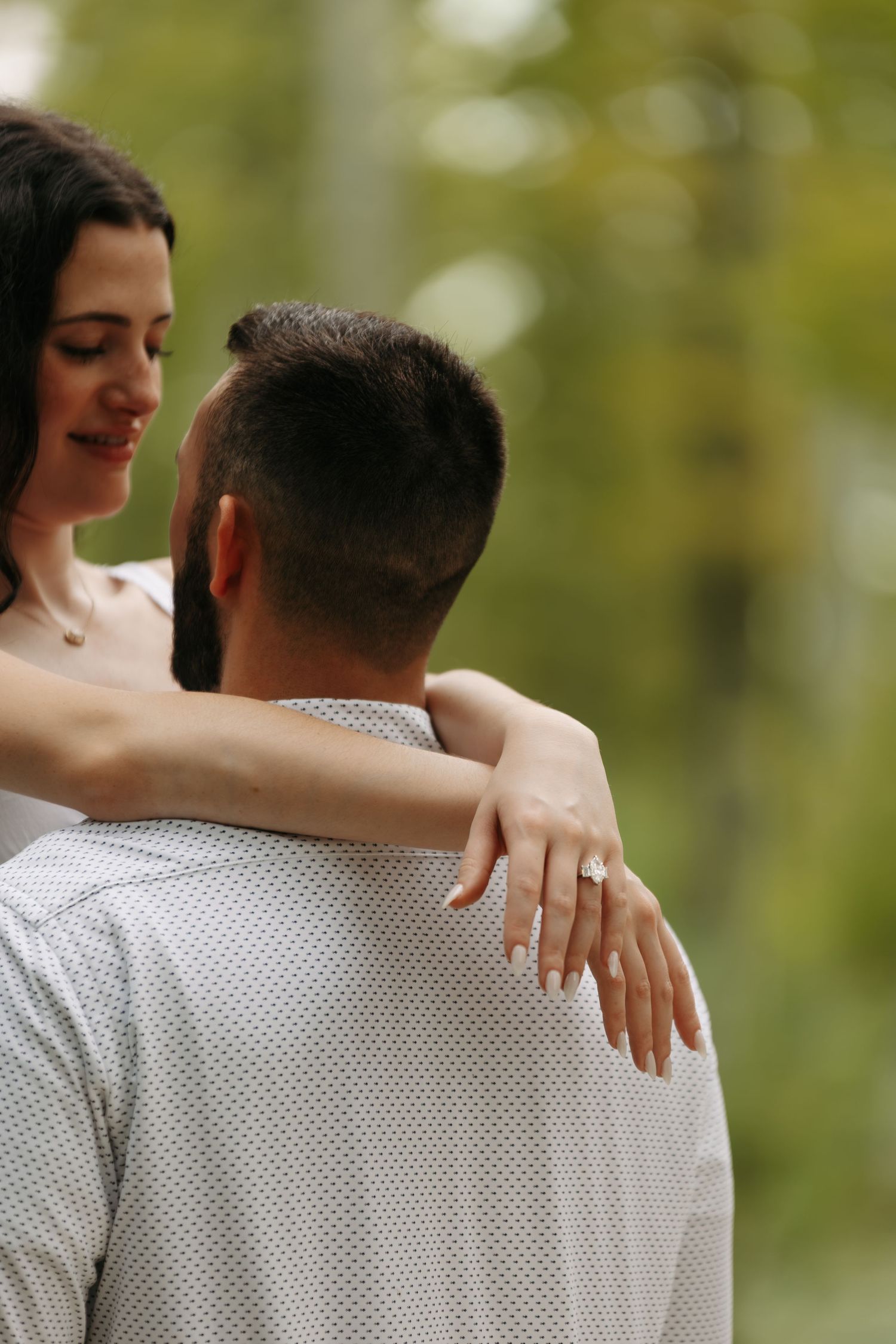 Engaged couple embracing outdoors, woman displaying diamond engagement ring over mans shoulder.