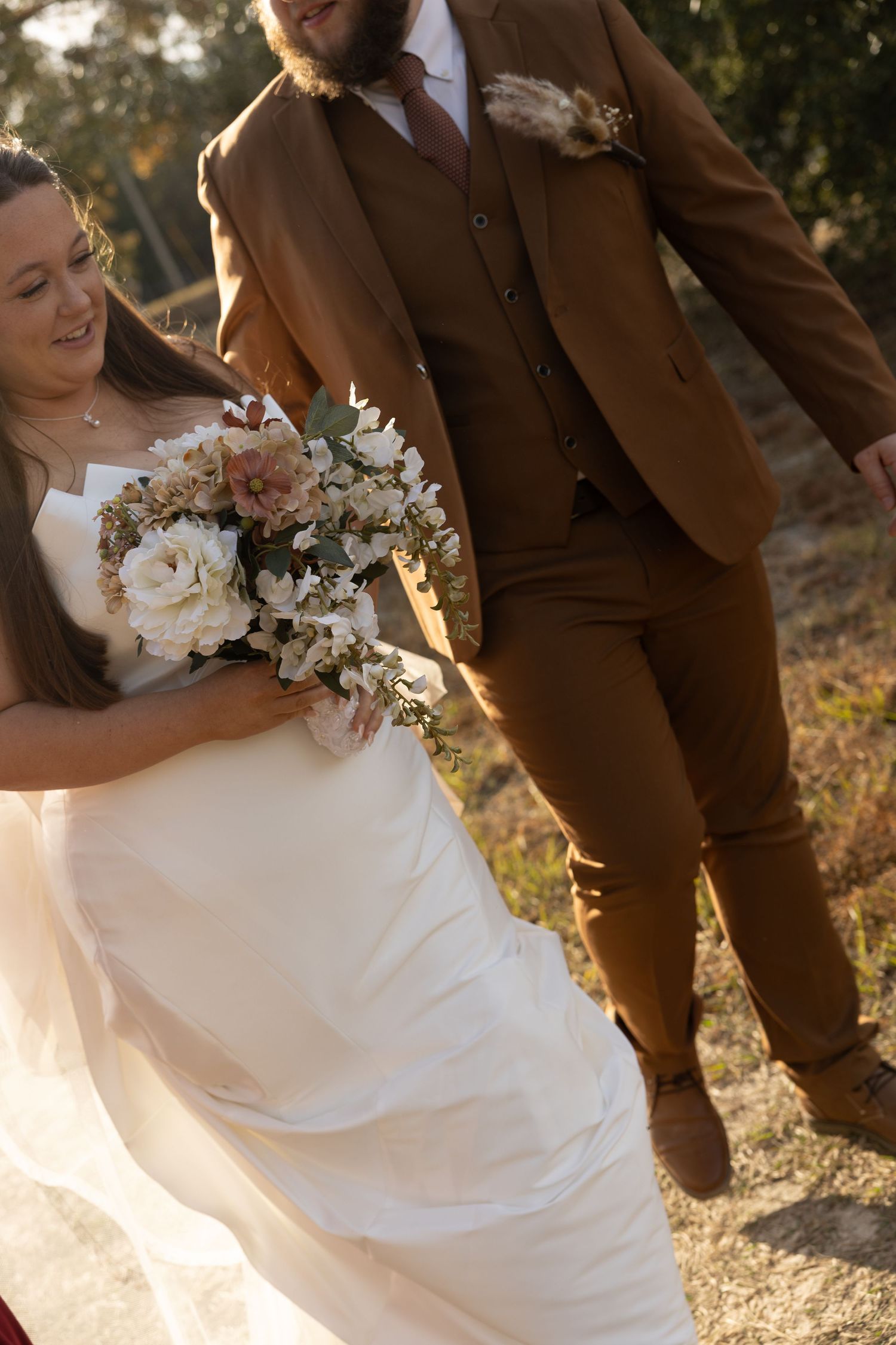 Bride in white gown holding floral bouquet walks with groom in brown three-piece suit outdoors in golden sunlight.