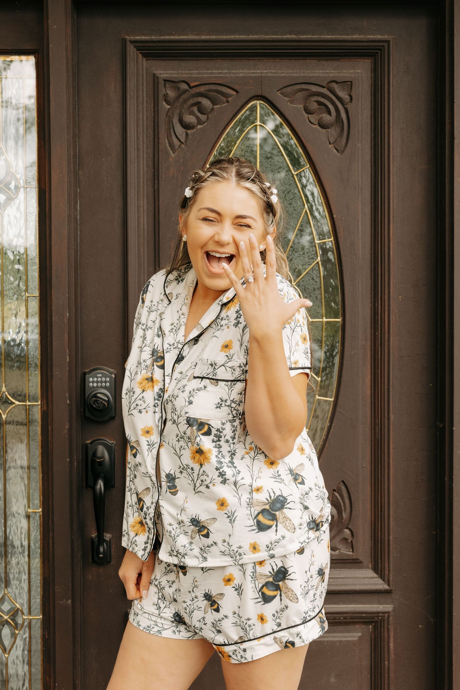 Joyful bride in floral bee-print pajamas showing off engagement ring while standing at ornate wooden door.