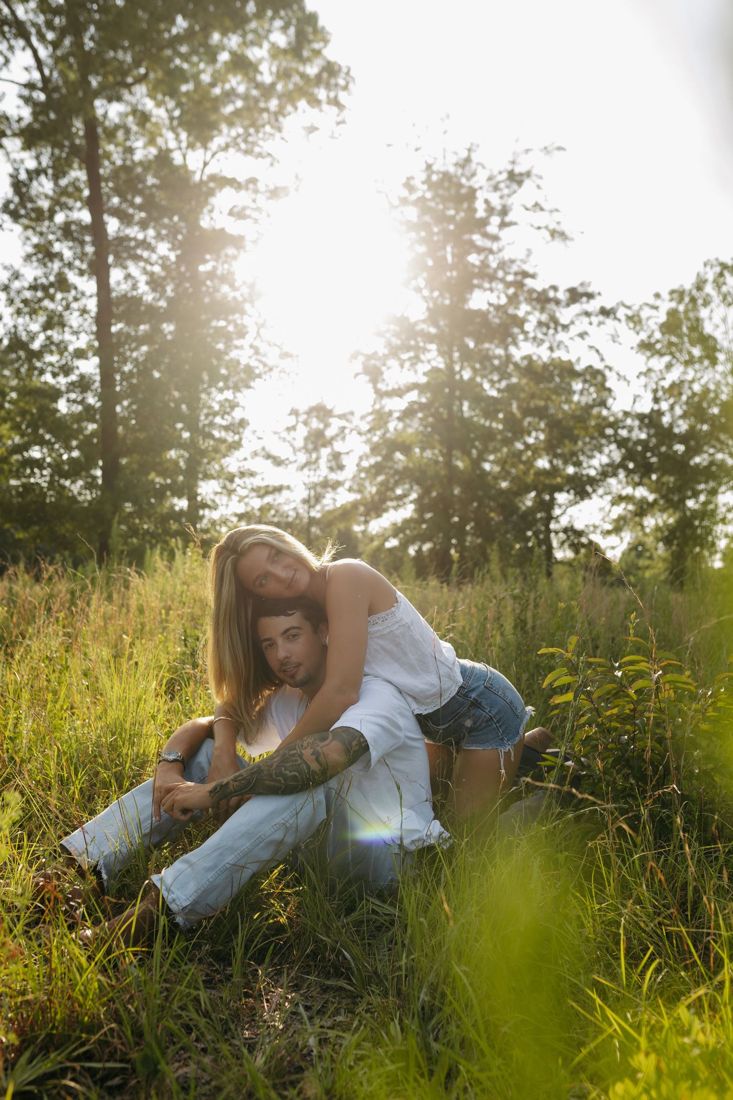 Couple sitting together in sunlit meadow with tall grass and trees, woman leaning over man affectionately outdoors.