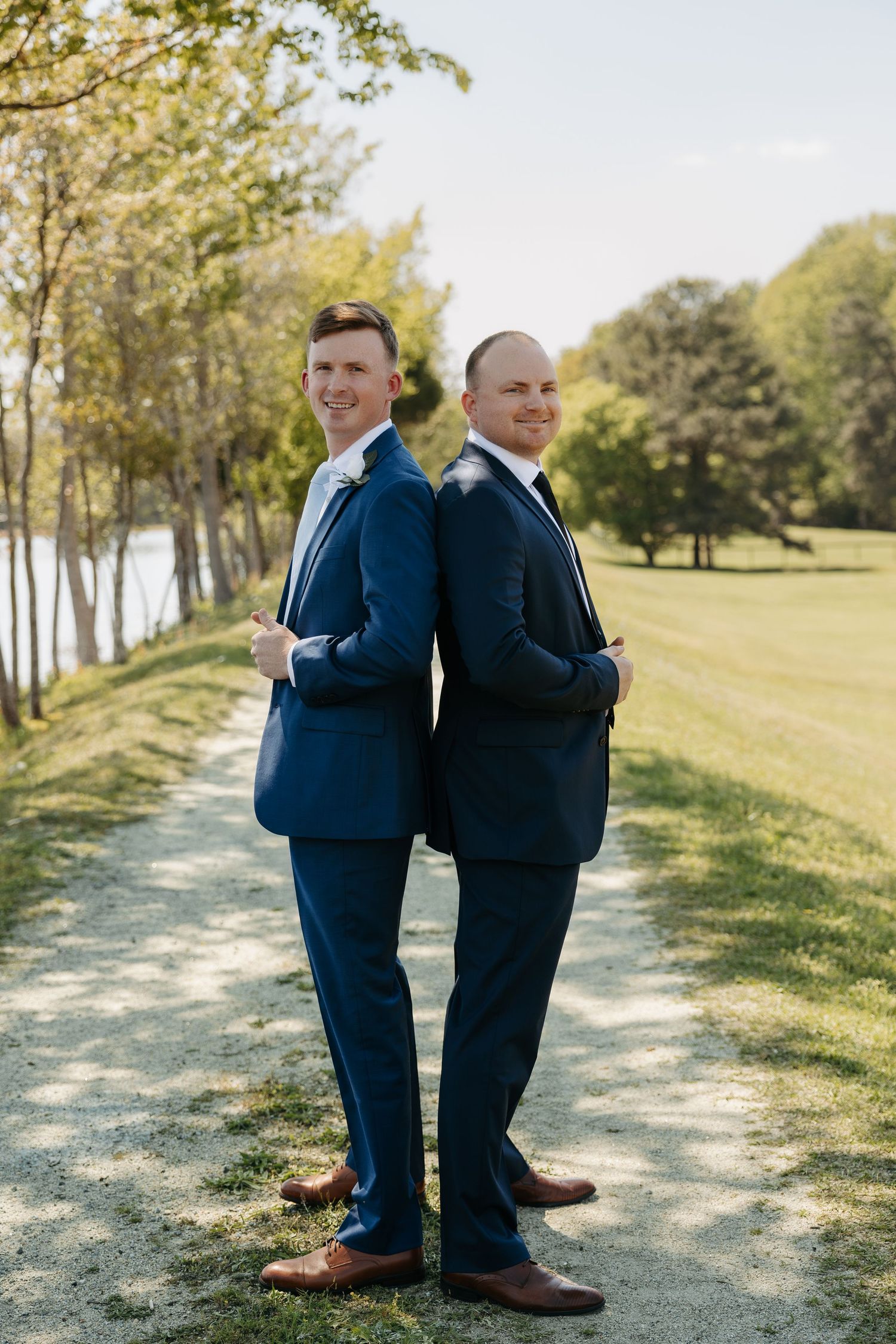 Two groomsmen in navy blue suits pose back-to-back on a scenic outdoor path surrounded by trees.