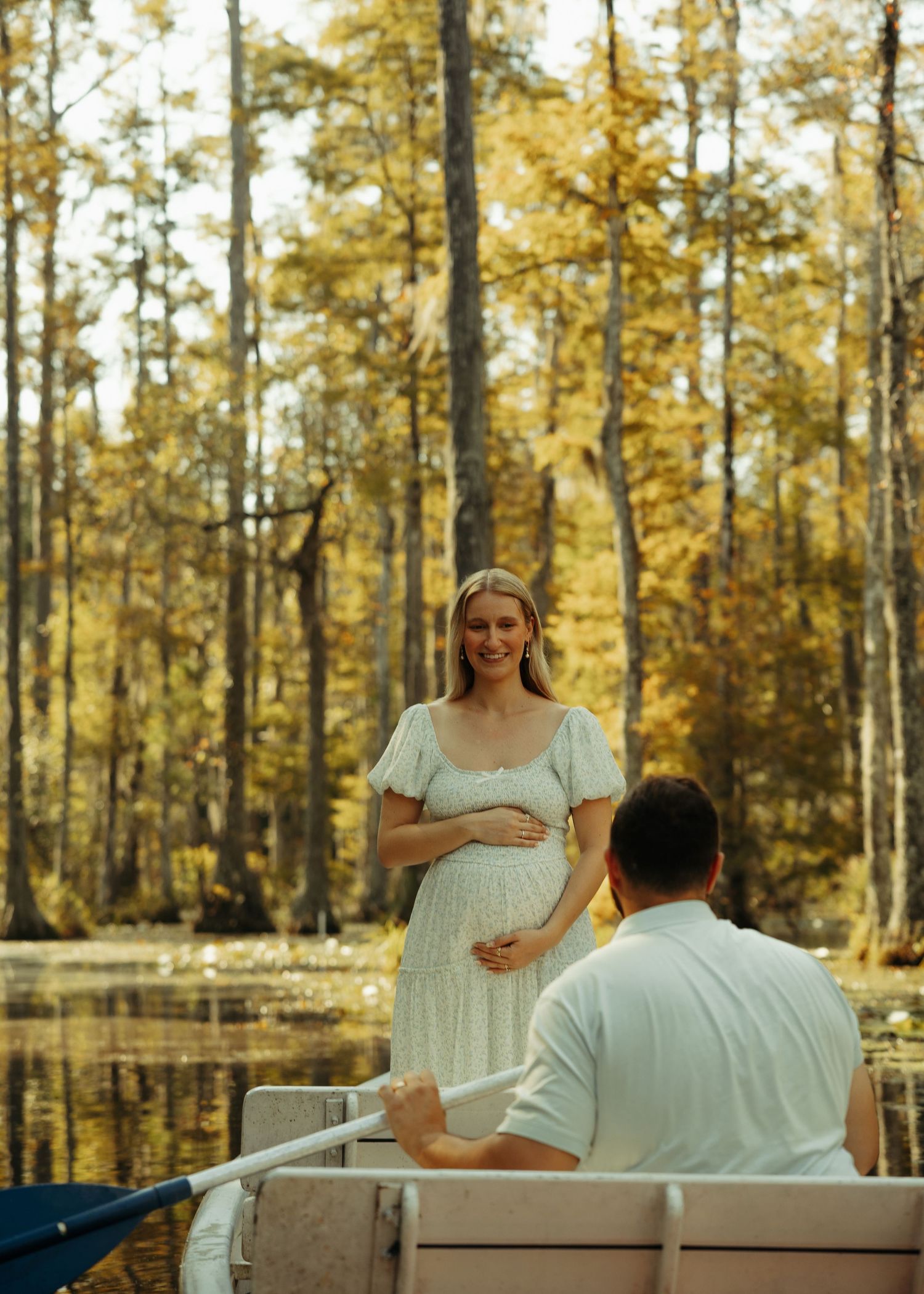 Pregnant woman in white dress on a boat, smiling at partner, surrounded by golden autumn trees reflected in calm water.