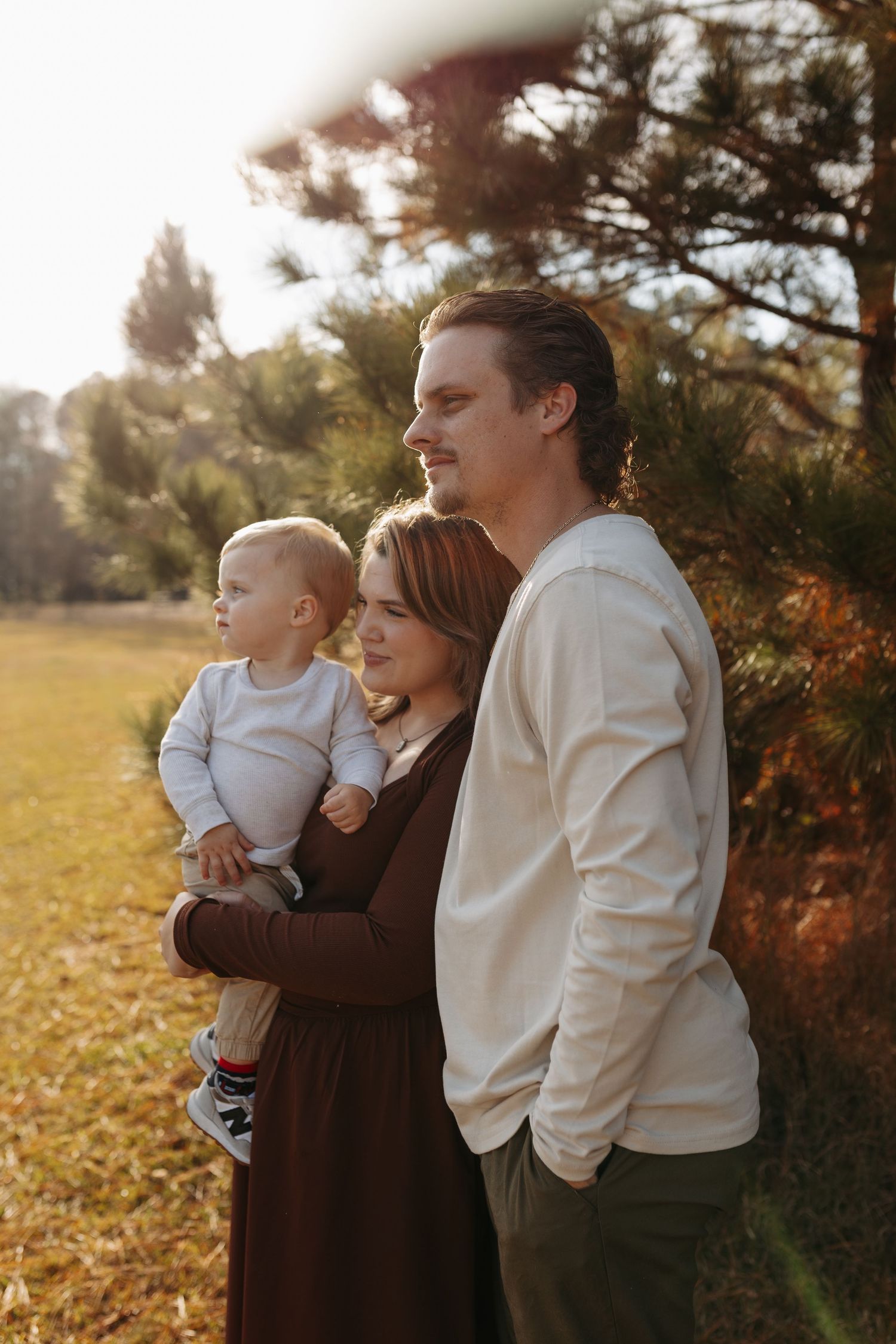 Young family with baby enjoying golden hour outdoors near pine trees in autumn park setting.