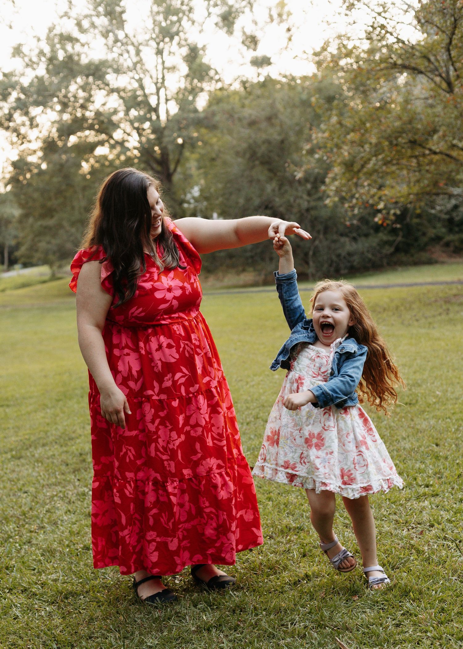 A woman in a red floral dress and a laughing girl in a white dress play together on a green lawn outdoors.