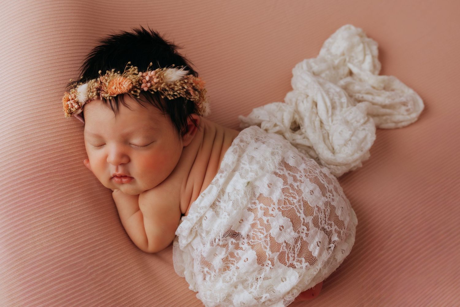 Sleeping newborn baby in delicate white lace wrap on pink background during professional photo session.